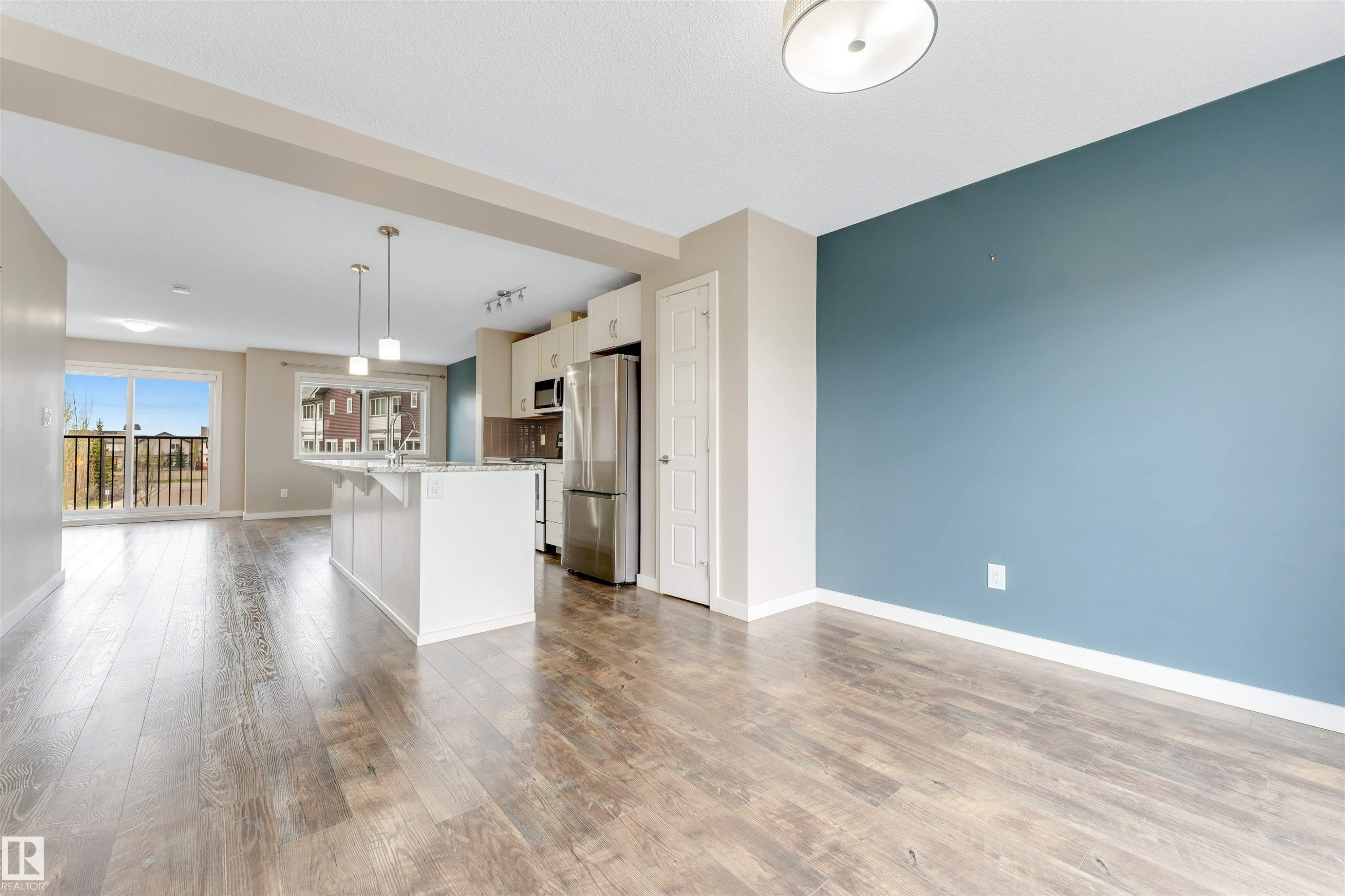 Expansive open-concept living area featuring wood-finish flooring, a contemporary kitchen island with pendant lighting, stainless steel appliances, and a sliding glass door leading to a balcony - 3340 15A Avenue, Edmonton, AB - Indoor Photo Showing Kitchen