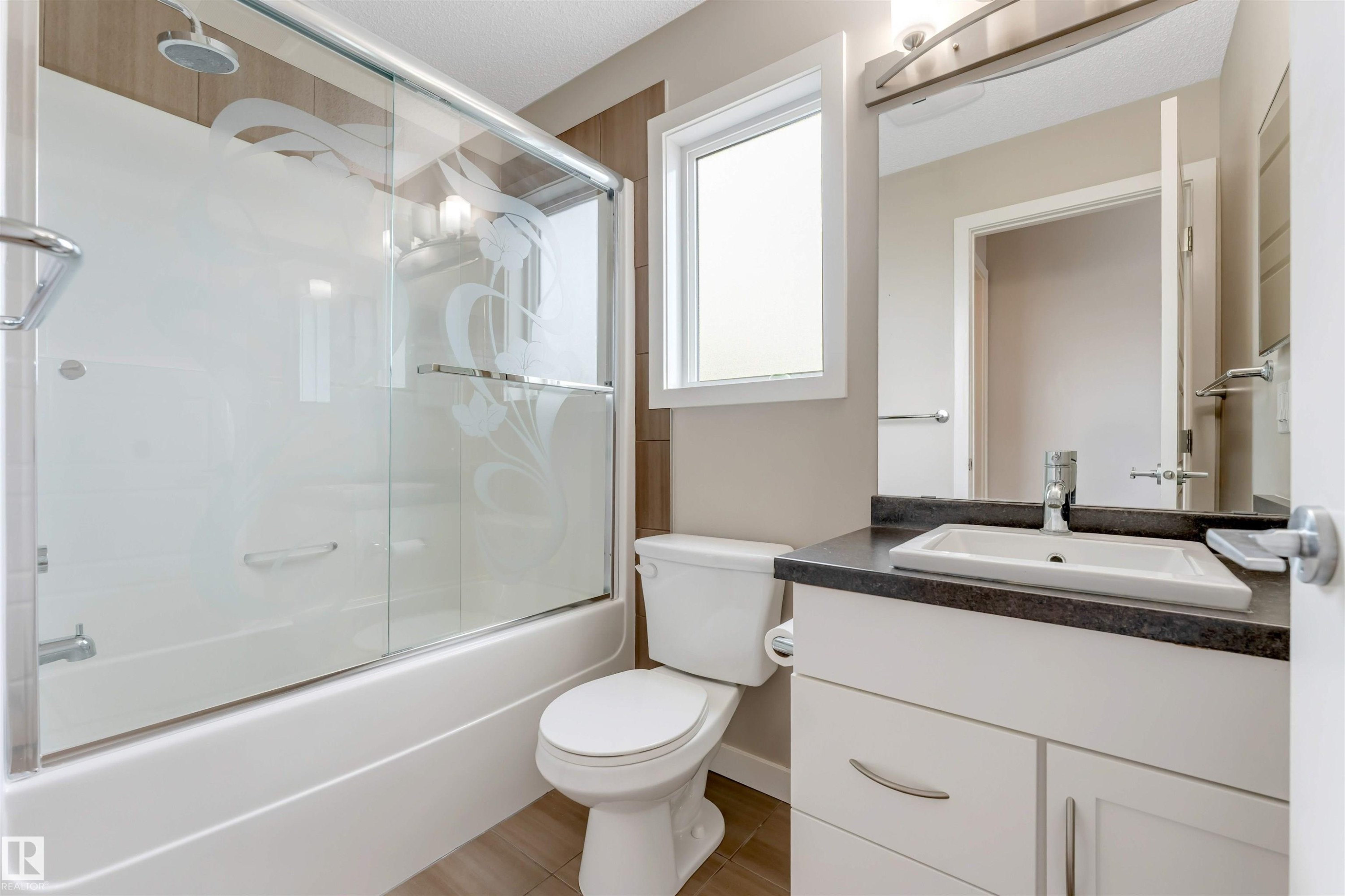 Bathroom featuring a bathtub with a clear glass sliding shower door, white toilet, white vanity with a dark countertop and undermount sink, and a rectangular window - 3340 15A Avenue, Edmonton, AB - Indoor Photo Showing Bathroom