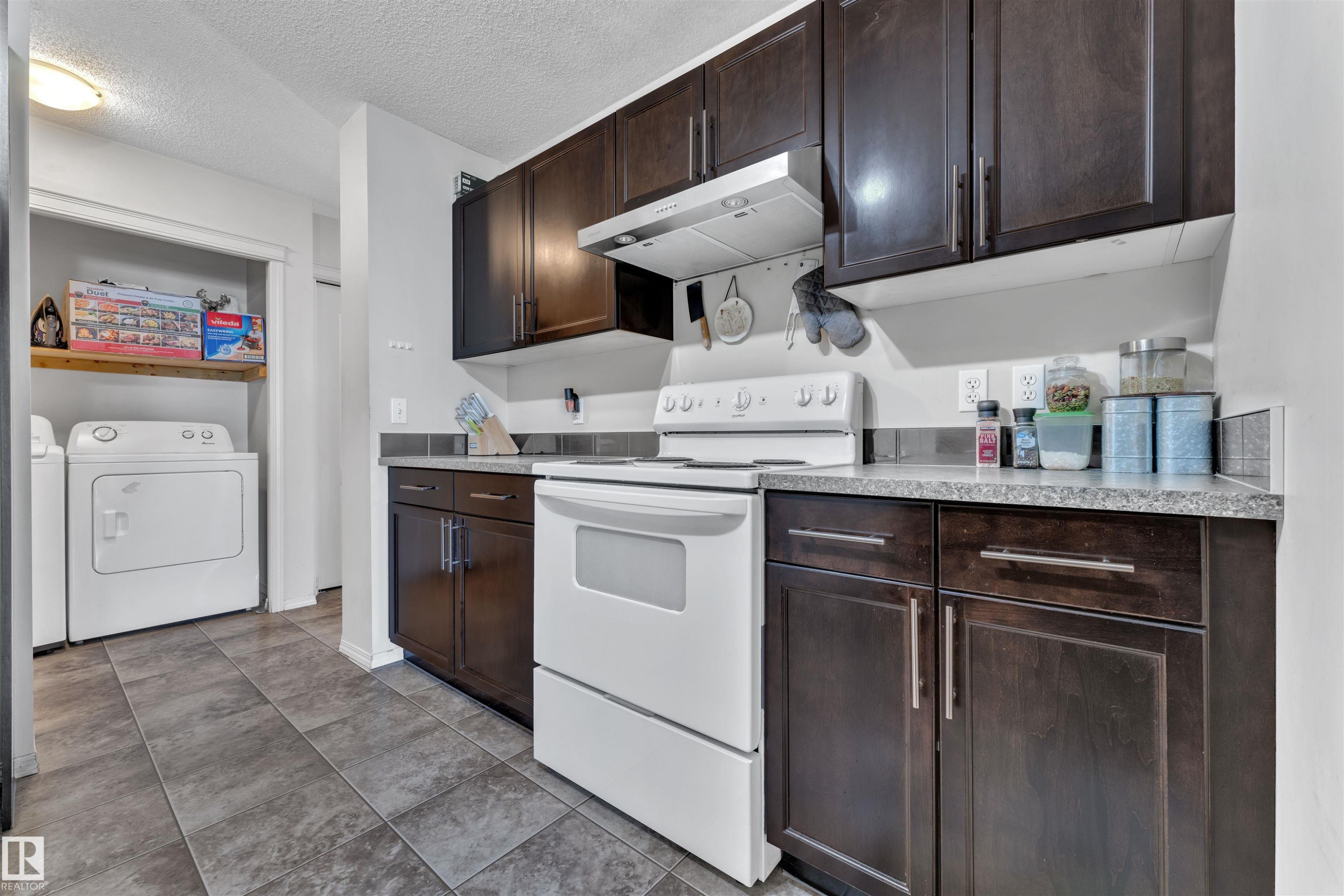 Kitchen featuring dark brown cabinets, white range with electric stovetop, under cabinet range hood, light countertops, and a textured ceiling - 6152 13 Avenue, Edmonton, AB - Indoor
