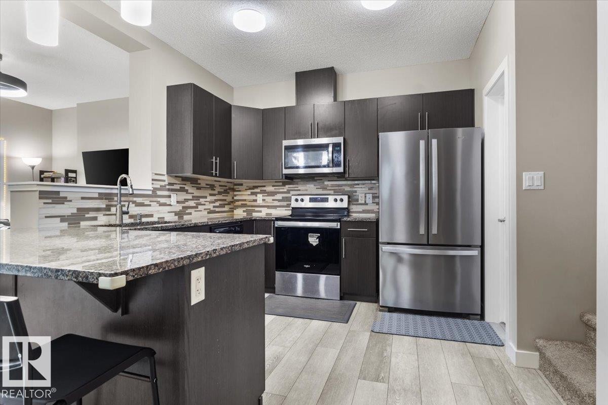 Modern kitchen featuring dark wood-finish cabinetry, stainless steel appliances, a grey patterned tile backsplash, a kitchen island with stone-finish countertop, and light wood-finish flooring - 236 39 Avenue, Edmonton, AB - Indoor Photo Showing Kitchen With Upgraded Kitchen