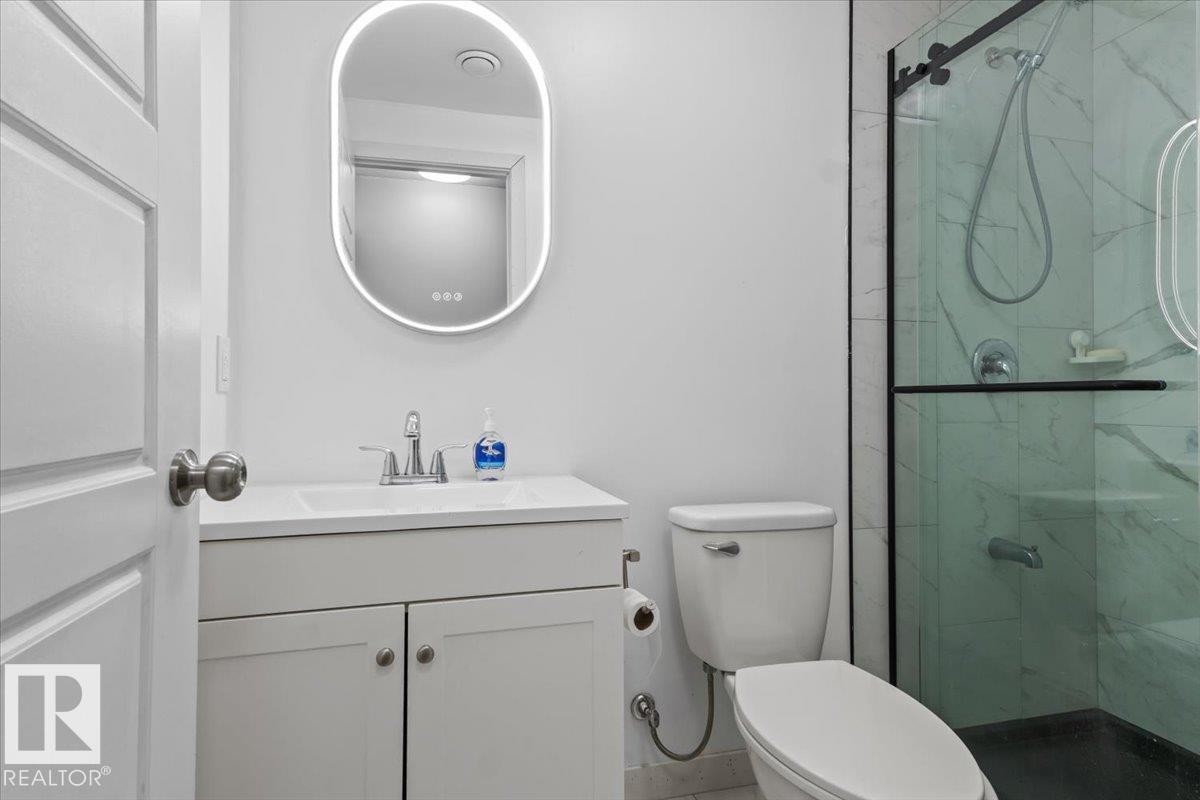 Modern bathroom featuring a contemporary vanity with an integrated sink, an illuminated oval mirror, a commode, and a glass-enclosed shower with white marble-patterned tile - 236 39 Avenue, Edmonton, AB - Indoor Photo Showing Bathroom