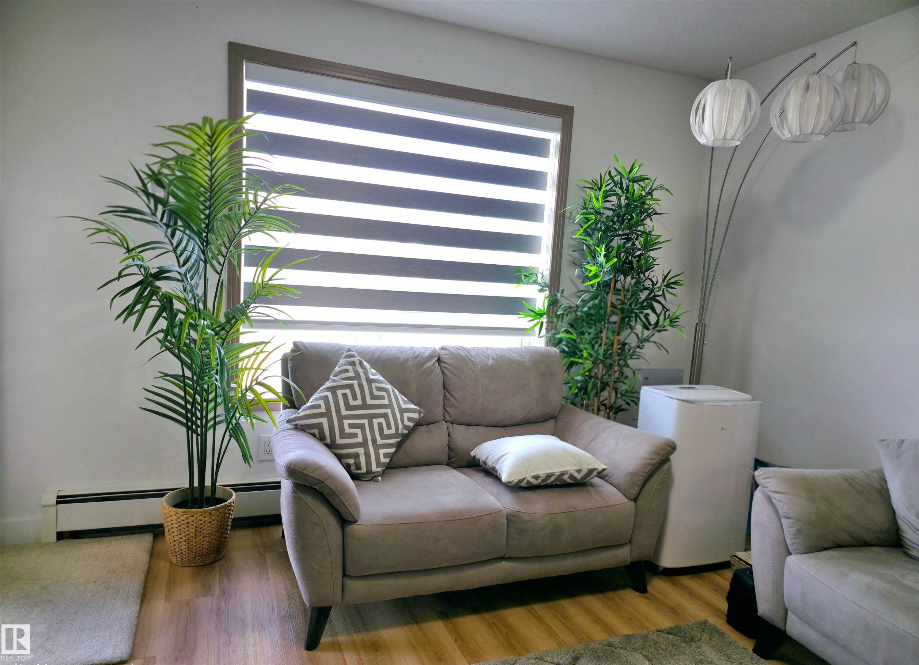 Inviting living space featuring wood-finish flooring, a baseboard heating unit, and a window with horizontal blinds - 210 5005 165 Avenue, Edmonton, AB - Indoor