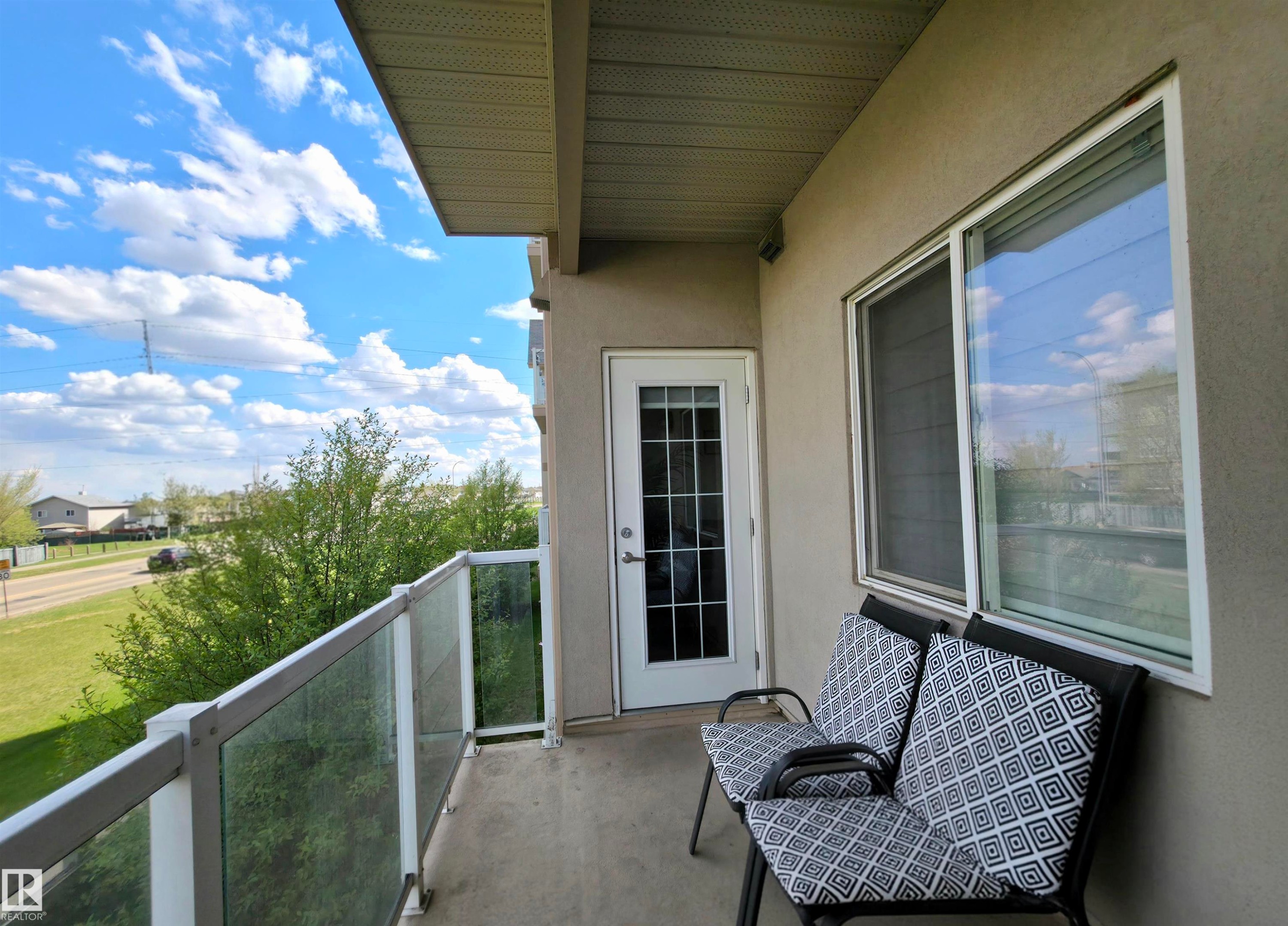 Private balcony featuring a glass railing system, stucco exterior, and a white framed window - 210 5005 165 Avenue, Edmonton, AB - Outdoor With Balcony With Exterior