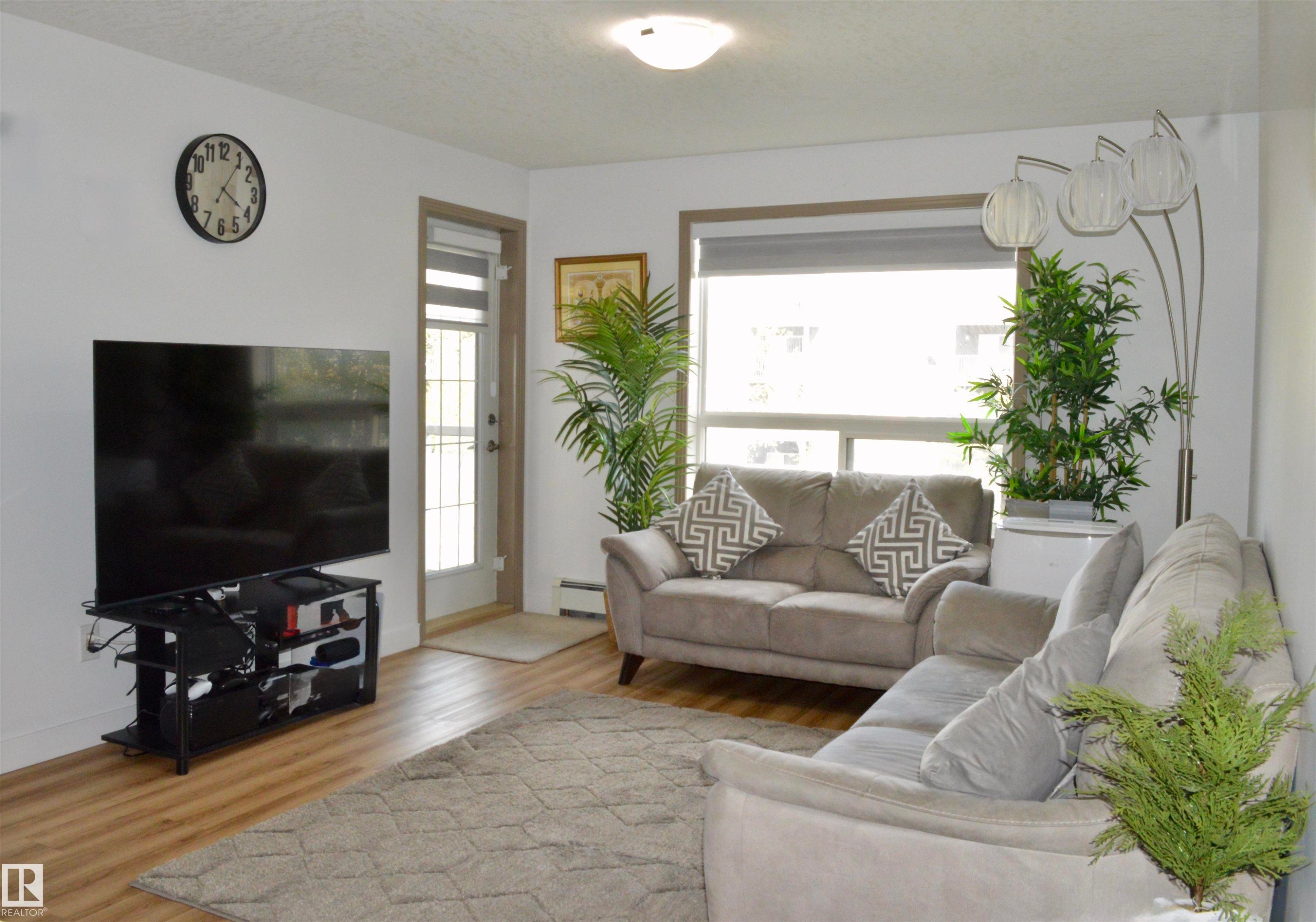 Living area featuring wood-finish flooring, white walls, and a large window with roller shades - 210 5005 165 Avenue, Edmonton, AB - Indoor Photo Showing Living Room