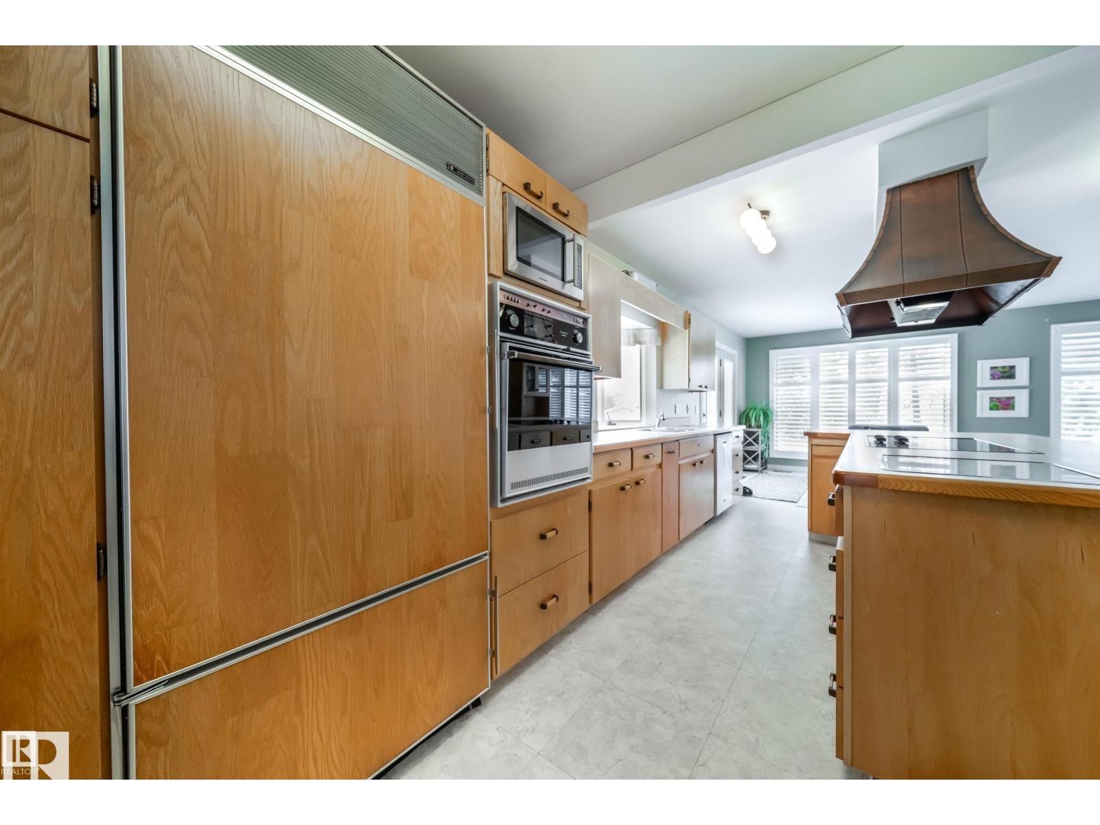 Kitchen featuring wood-panel appliances, built-in wall oven and microwave, an island with cooktop, a distinctive copper-toned range hood, and white plantation shutters - 10231 46 Street, Edmonton, AB
