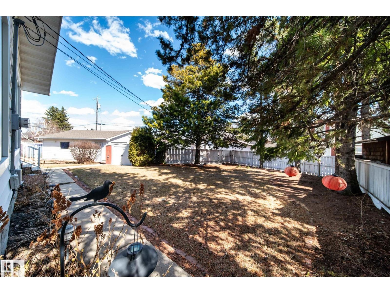 Expansive backyard featuring mature trees, a concrete pathway, a detached garage with an overhead door, and a white picket fence - 10231 46 Street, Edmonton, AB