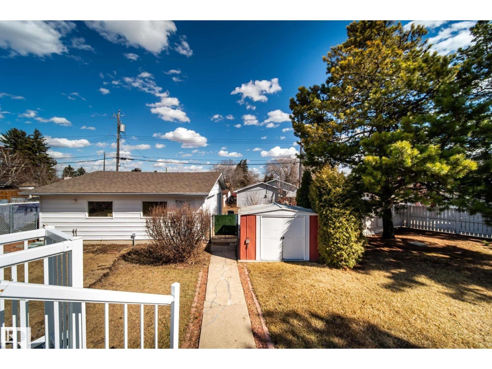 Spacious backyard featuring a white picket fence, large mature tree, a red and white storage shed, and a concrete pathway leading to the main structure - 10231 46 Street, Edmonton, AB