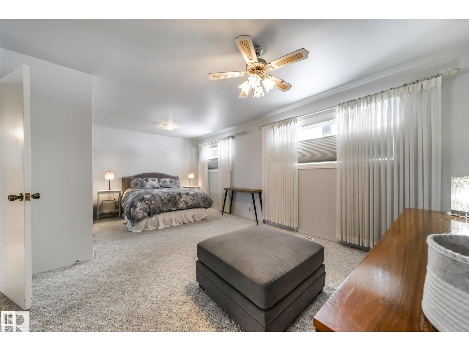 Carpeted bedroom featuring multiple windows with privacy blinds and sheer curtains, a ceiling fan with integrated lighting, and a neutral white wall finish - 10231 46 Street, Edmonton, AB