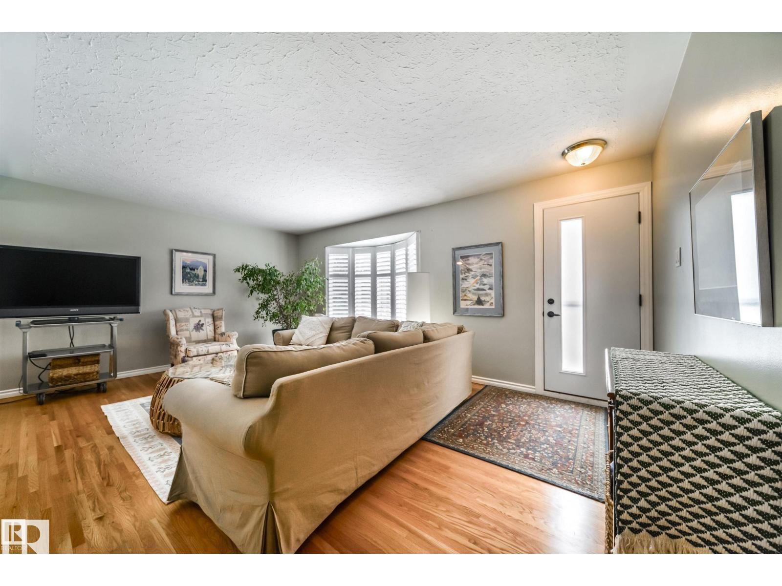 Living area featuring wood-finish flooring, a bay window with plantation shutters, a white entry door with frosted glass insert, and a flush-mount ceiling light fixture - 10231 46 Street, Edmonton, AB