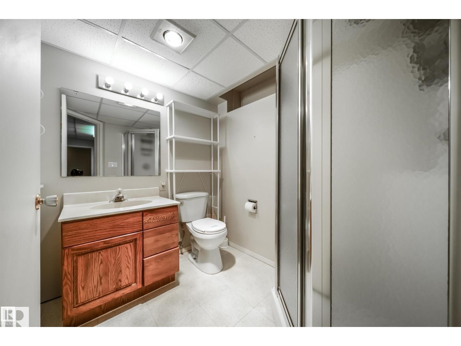 Full bathroom featuring a wood-finish vanity with an integrated sink, a wall-mounted mirror with overhead lighting, a toilet, and a stand-up shower with frosted glass doors - 10231 46 Street, Edmonton, AB