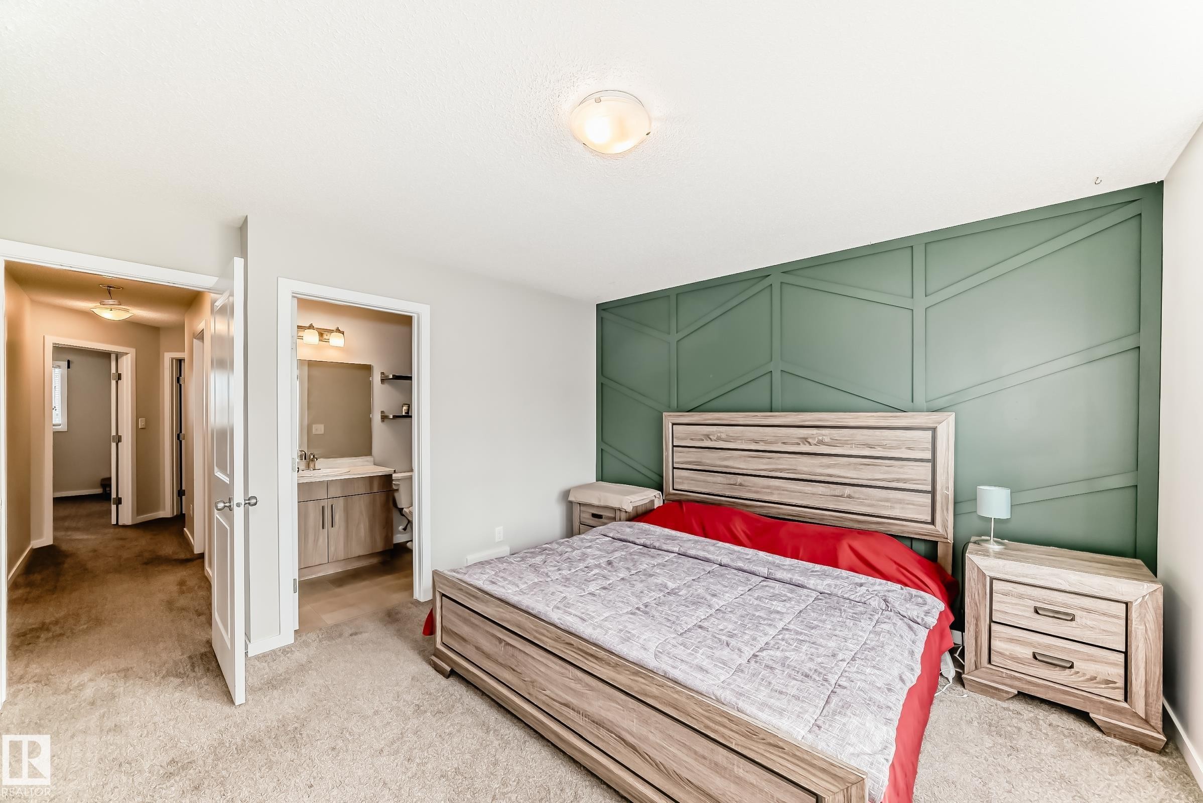 Carpeted bedroom featuring a decorative accent wall with geometric molding, a ceiling-mounted light fixture, and a doorway to a bathroom with a vanity and wall-mounted lighting - 328 Watt Boulevard, Edmonton, AB - Indoor Photo Showing Bedroom