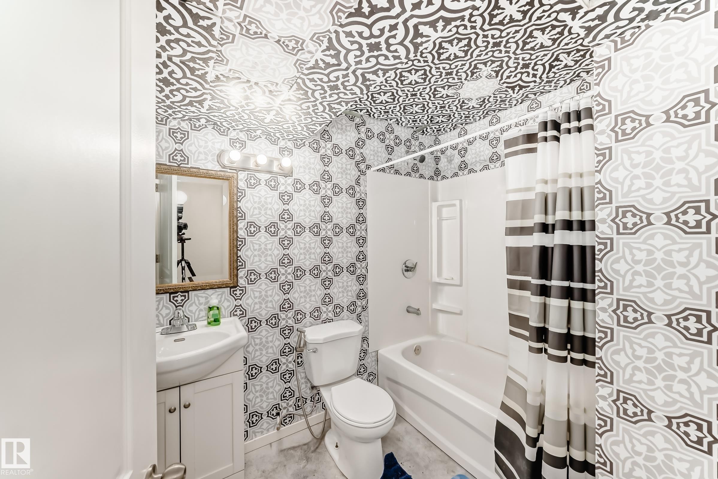 Bathroom featuring patterned wall and ceiling accents, a white vanity with integrated sink, a framed mirror, a four-bulb vanity light fixture, and a tub-shower combination with white surround - 328 Watt Boulevard, Edmonton, AB - Indoor Photo Showing Bathroom