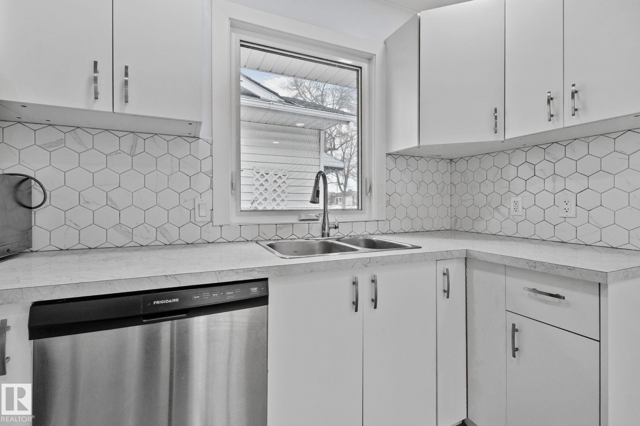 Kitchen featuring a stainless steel double basin sink, white cabinetry with silver hardware, hexagon tile backsplash, light-toned countertops, and a built-in stainless steel dishwasher - 12002 37 Street, Edmonton, AB - Indoor Photo Showing Kitchen With Double Sink With Upgraded Kitchen