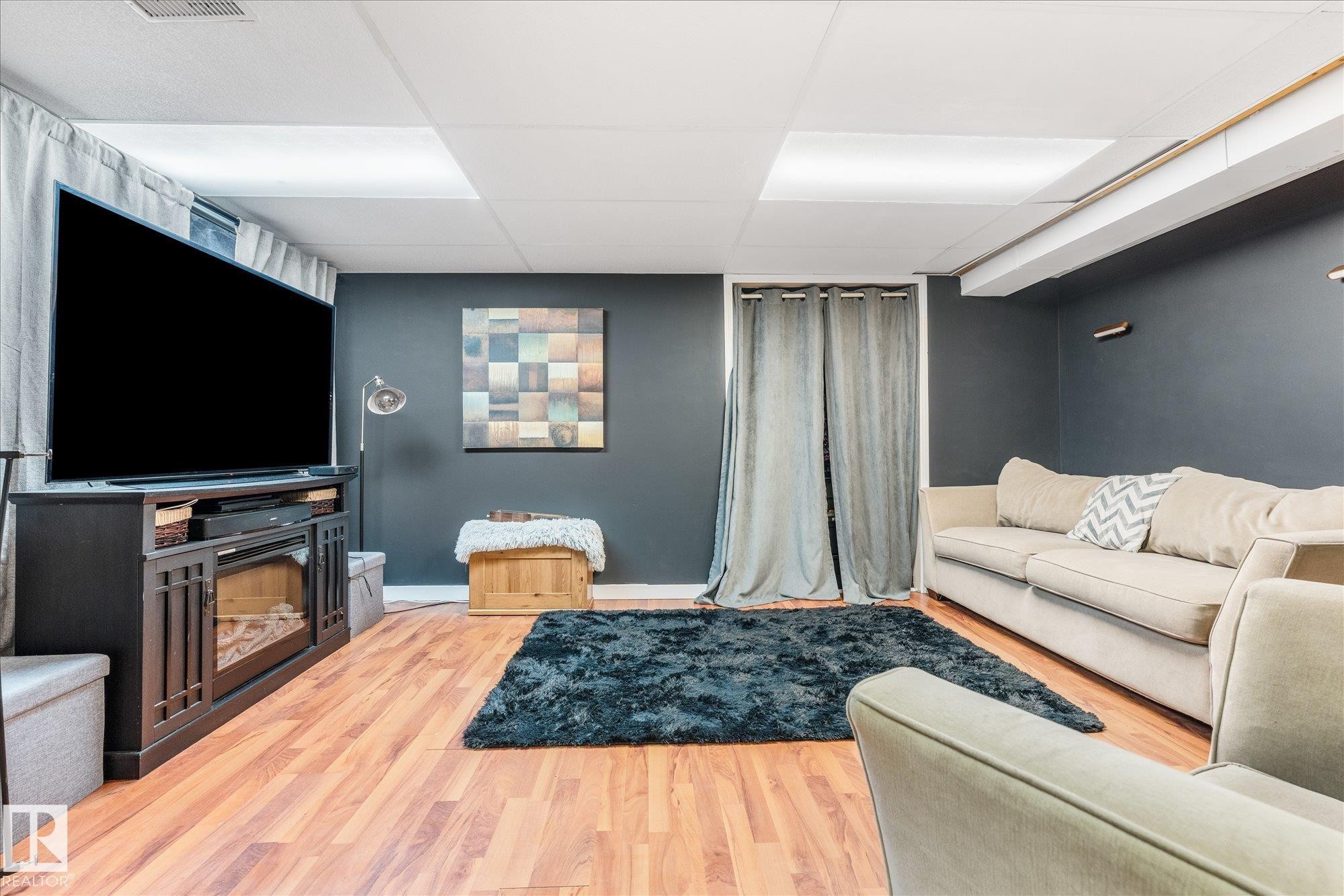 Recreation room featuring wood-finish flooring, dark accent walls, and a drop ceiling with integrated lighting panels - 4759 48 Street, Onoway, AB - Indoor With Fireplace