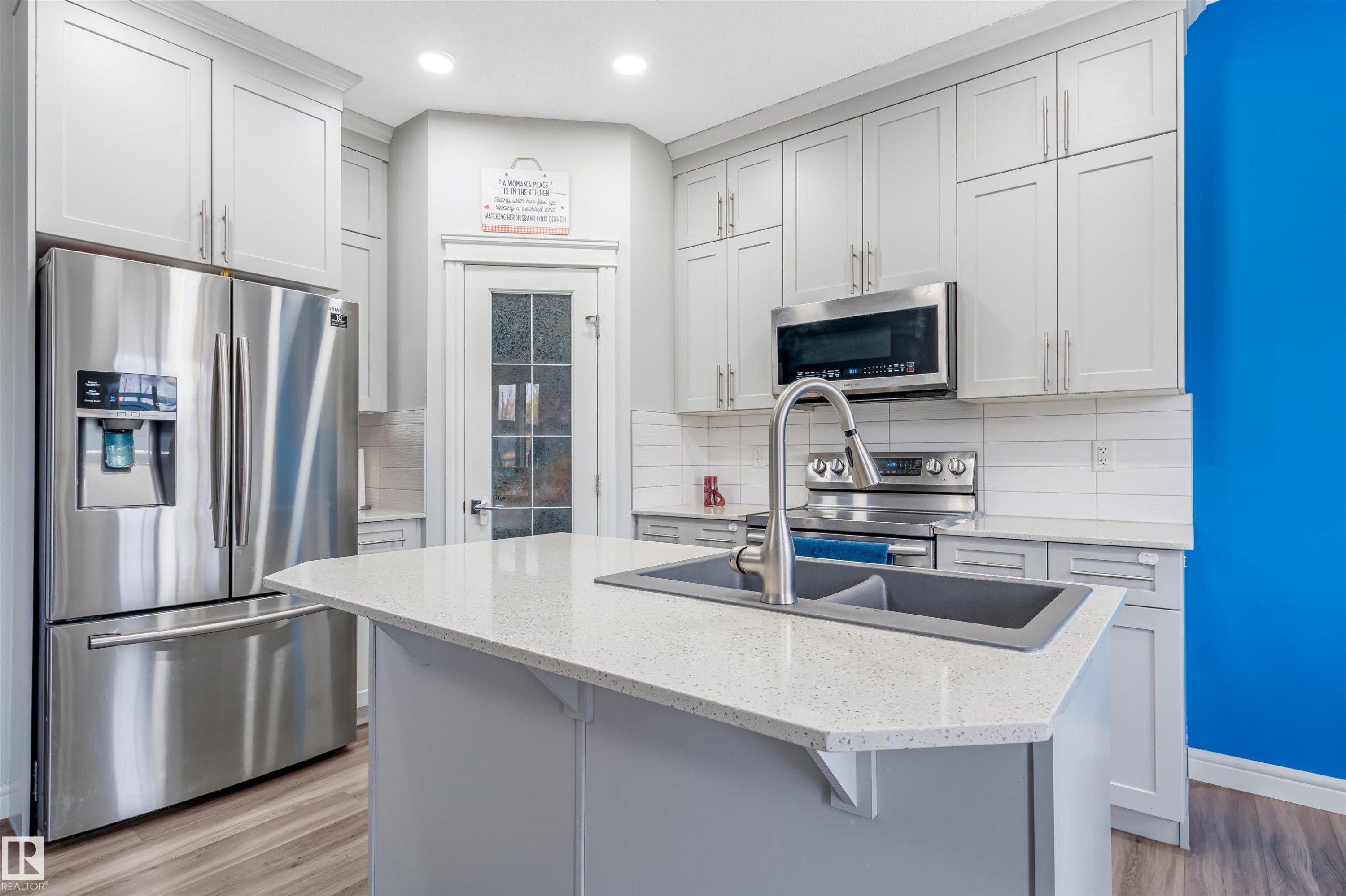 Kitchen featuring a spacious island with an undermount sink, light-toned countertops, contemporary light gray cabinetry, stainless steel appliances, and wood-finish flooring - 1024 Daniels Loop, Edmonton, AB - Indoor Photo Showing Kitchen With Double Sink With Upgraded Kitchen