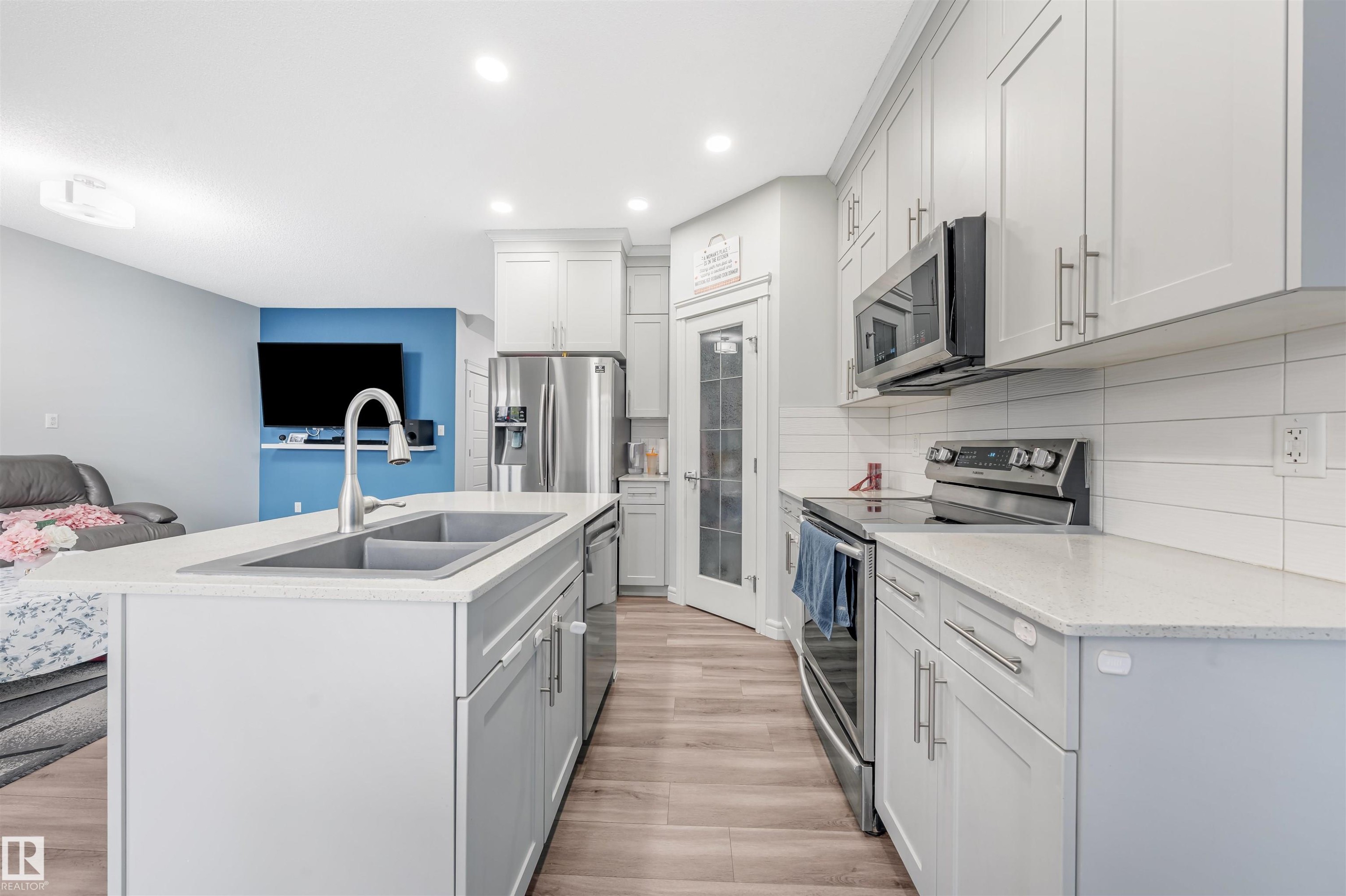 Modern kitchen featuring light gray cabinetry, stainless steel appliances, light-toned countertops, wood-finish flooring, and a central island with a double basin sink - 1024 Daniels Loop, Edmonton, AB - Indoor Photo Showing Kitchen With Double Sink With Upgraded Kitchen