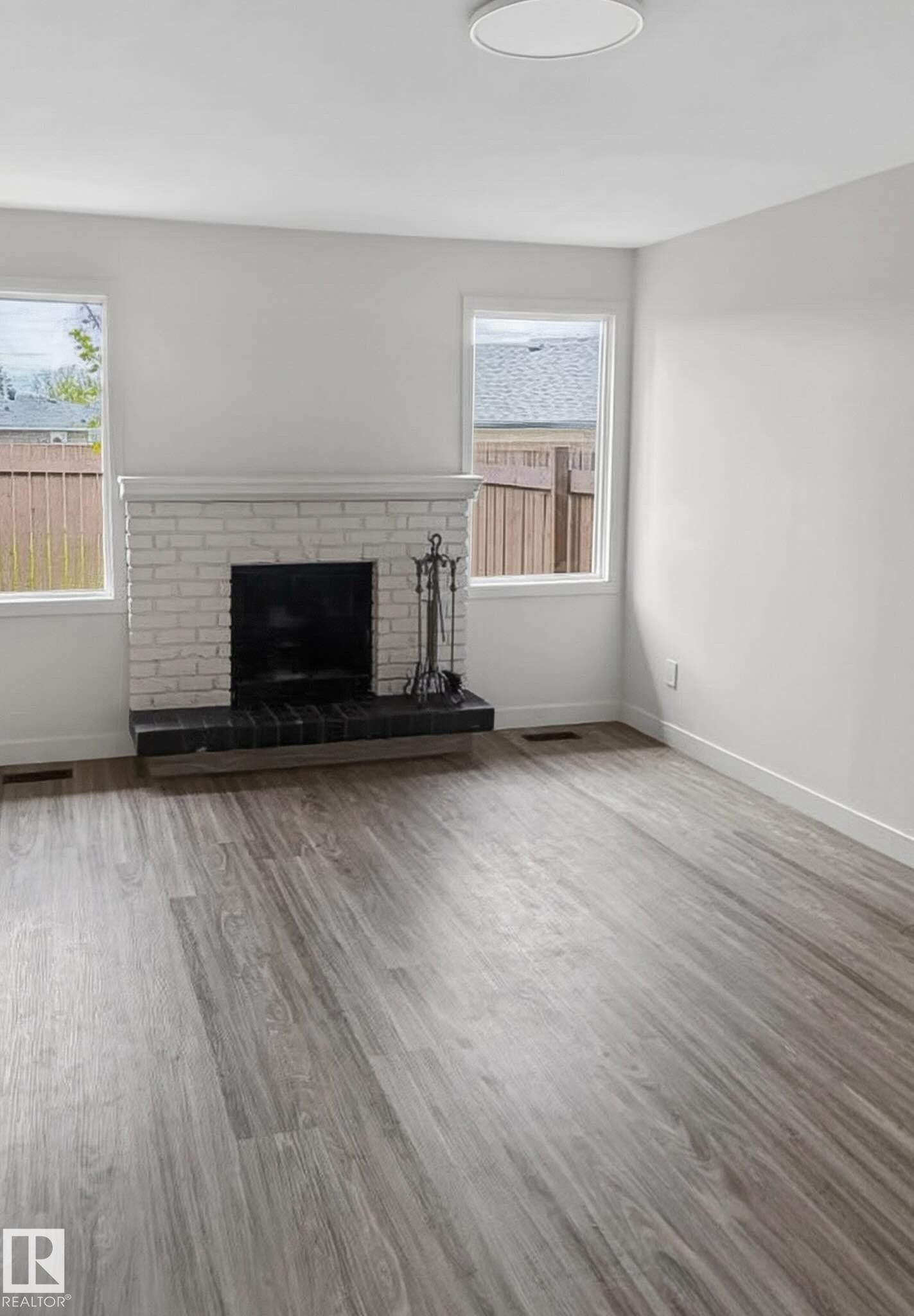 Living area featuring a white-painted brick fireplace with a dark hearth, wood-finish flooring, two large windows, and a flush-mount ceiling light - 13833 24 Street W, Edmonton, AB - Indoor Photo Showing Living Room With Fireplace