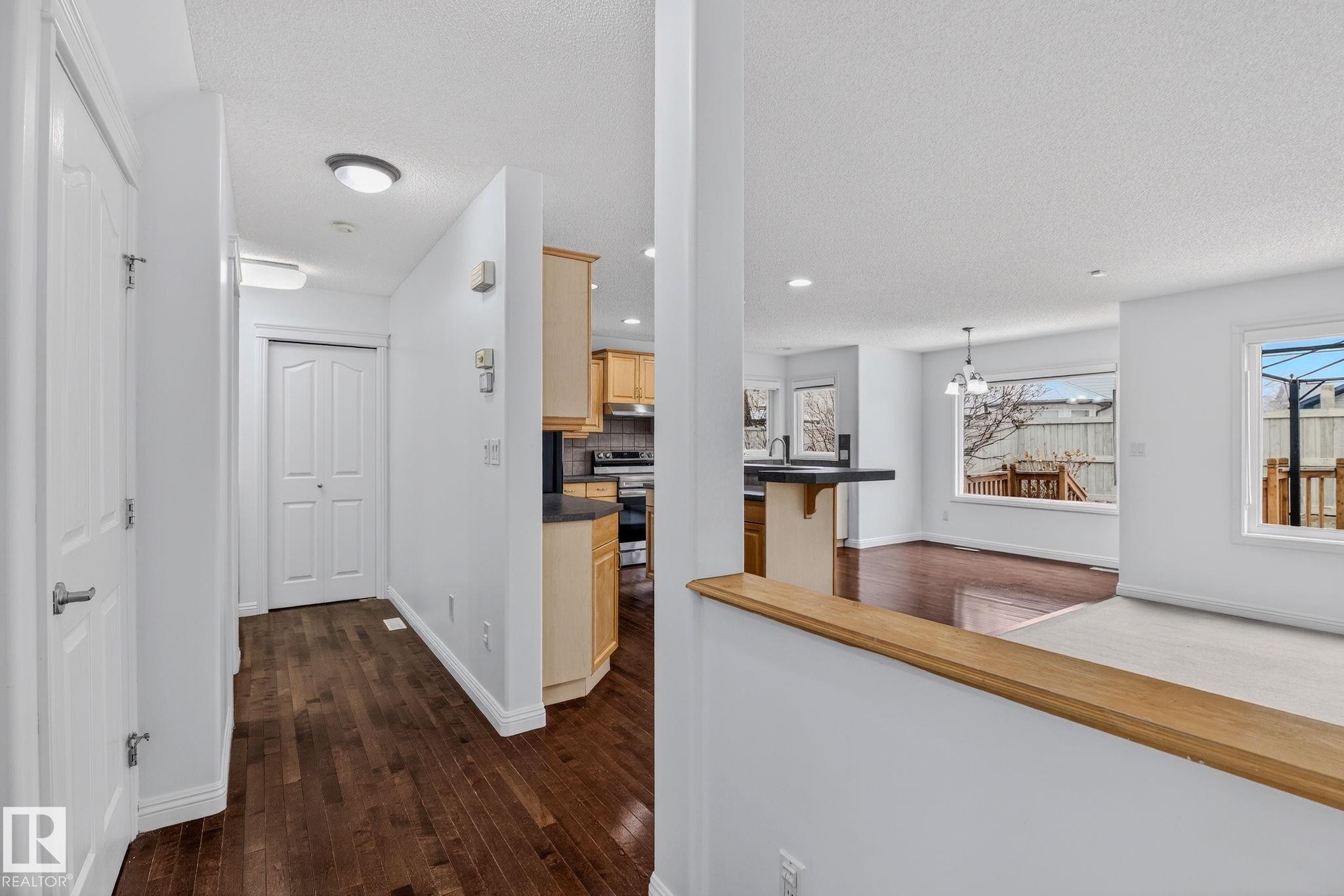 Entryway with dark wood-finish flooring, white walls, and a built-in closet, opening to a kitchen with light wood cabinetry and a peninsula countertop, and a living area featuring large windows - 978 Rutherford Road Sw, Edmonton, AB - Indoor Photo Showing Other Room