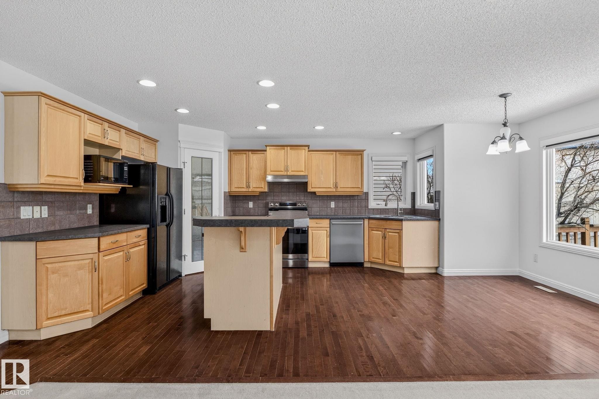 Open-concept kitchen with wood-finish flooring, light wood cabinetry, dark countertops, and stainless-steel appliances - 978 Rutherford Road Sw, Edmonton, AB - Indoor Photo Showing Kitchen