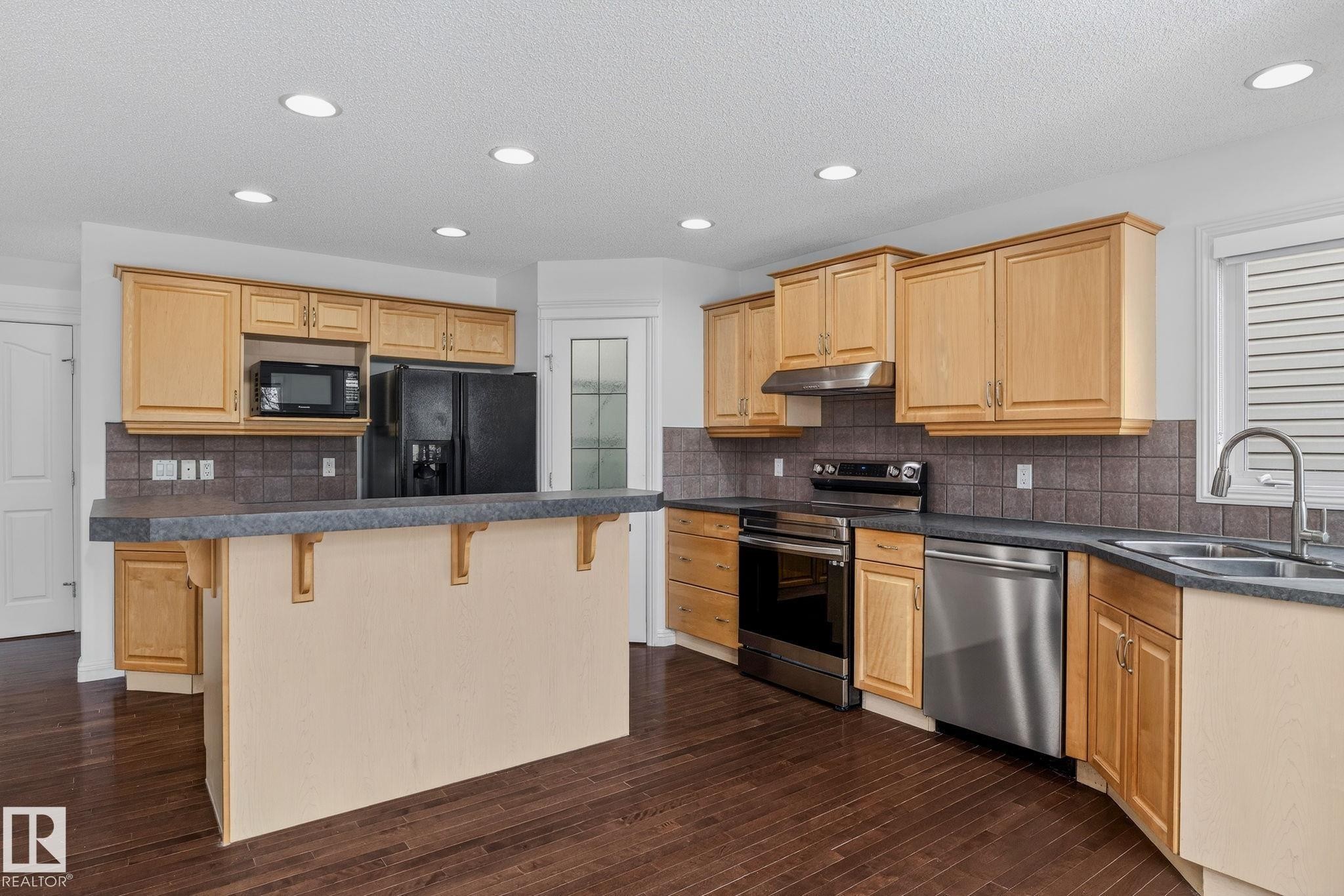 Kitchen featuring wood-finish flooring, recessed lighting, light wood cabinetry, a breakfast bar, and stainless steel appliances - 978 Rutherford Road Sw, Edmonton, AB - Indoor Photo Showing Kitchen With Double Sink