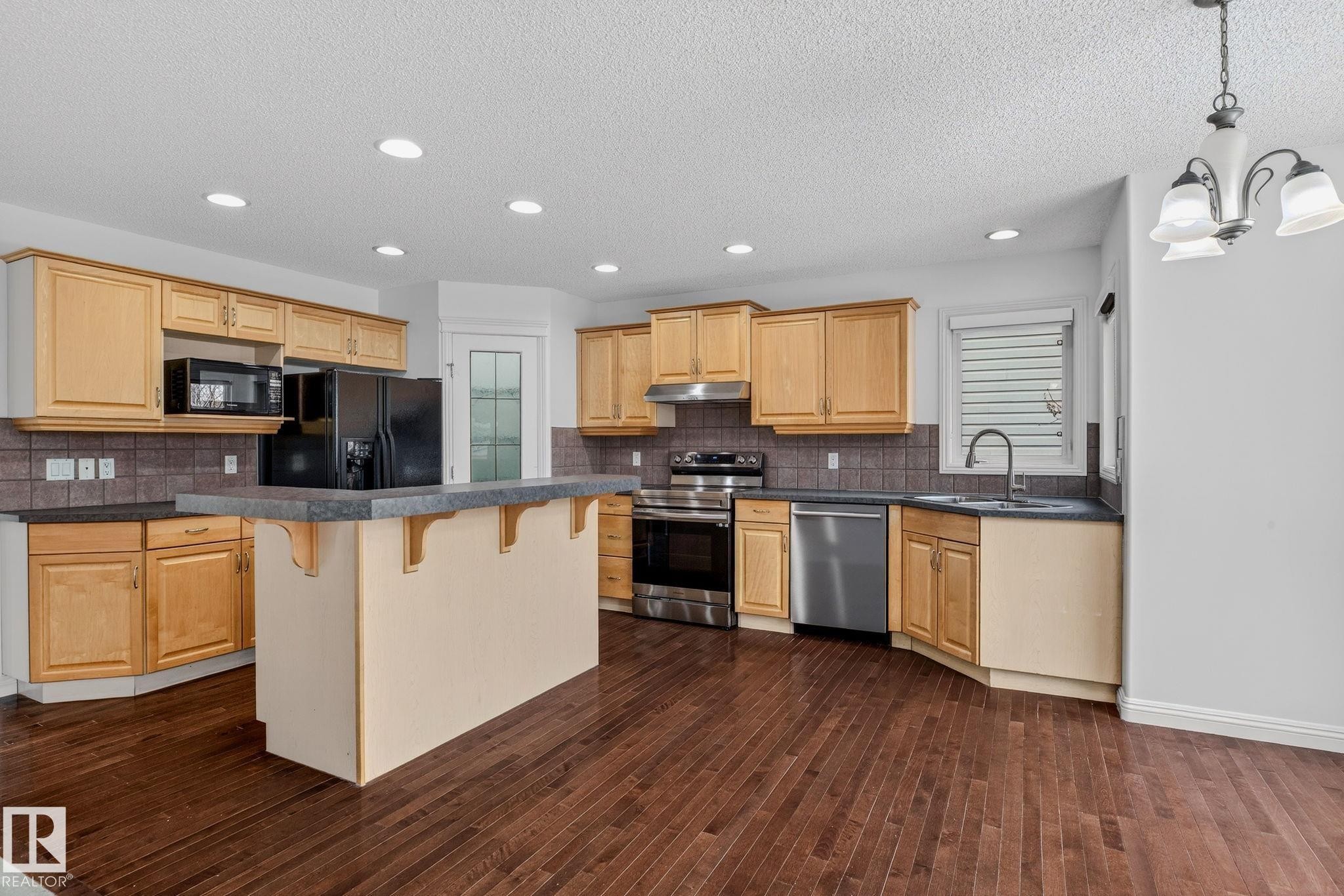 Spacious kitchen featuring wood cabinetry, stainless steel appliances, a center island with seating, a tile backsplash, and dark wood-finish flooring - 978 Rutherford Road Sw, Edmonton, AB - Indoor Photo Showing Kitchen With Double Sink