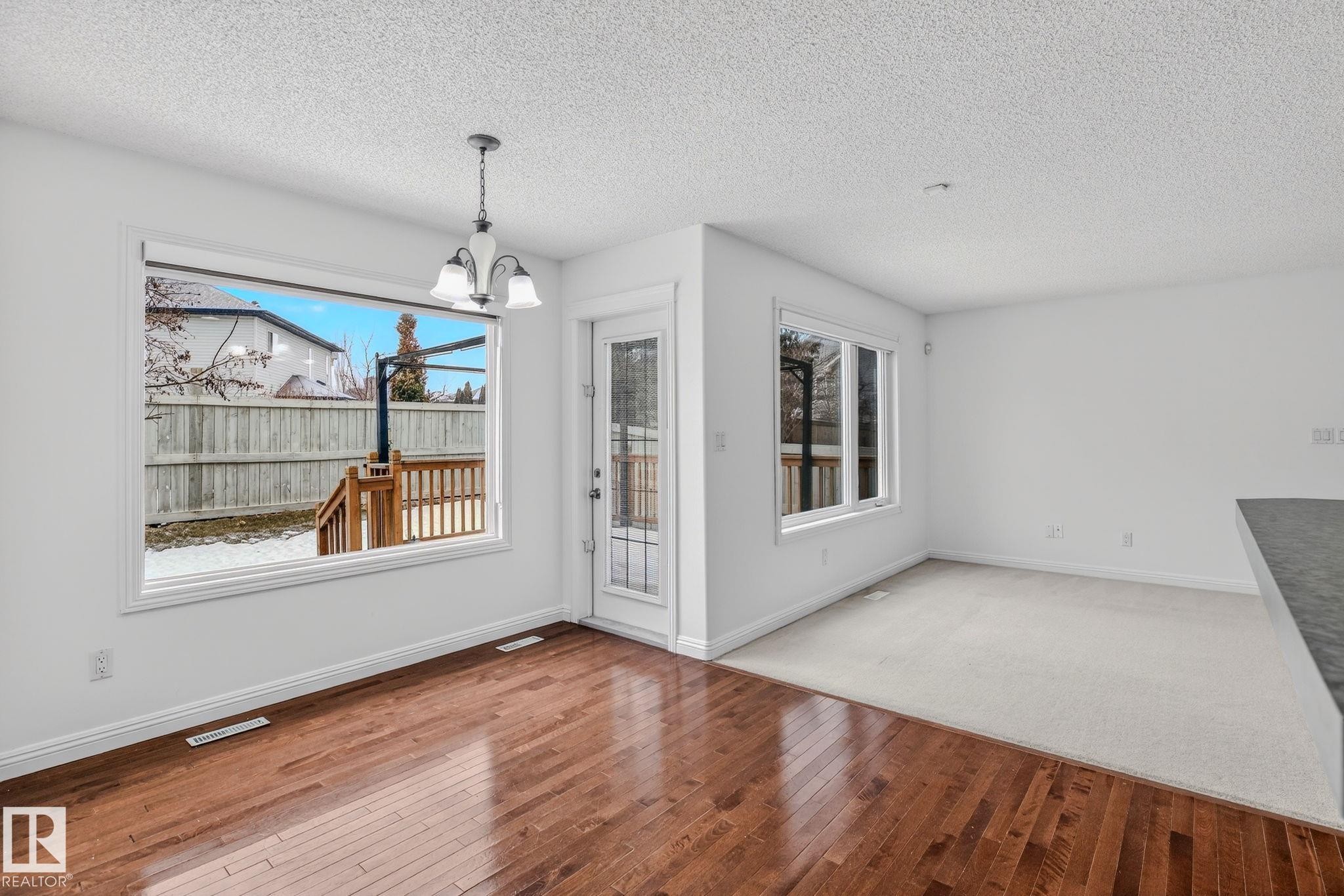 Bright dining area featuring a prominent window, a glass-paned door opening to an outdoor deck, a hanging chandelier, rich wood-finish flooring, and white baseboards - 978 Rutherford Road Sw, Edmonton, AB - Indoor