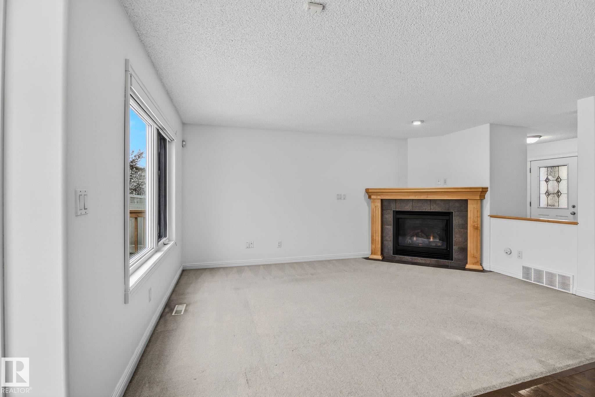 Spacious living area featuring a gas fireplace with a wood mantel and stone-tile surround, light-toned carpeting, and a large window providing ample natural light - 978 Rutherford Road Sw, Edmonton, AB - Indoor Photo Showing Living Room With Fireplace