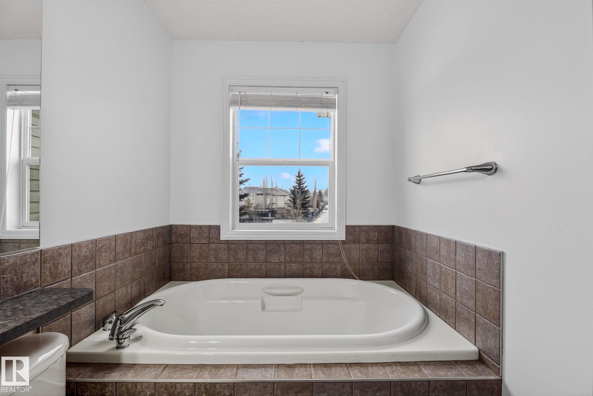 Built-in soaking tub with chrome fixtures, surrounded by brown square tilework, and a window with blinds - 978 Rutherford Road Sw, Edmonton, AB - Indoor Photo Showing Bathroom