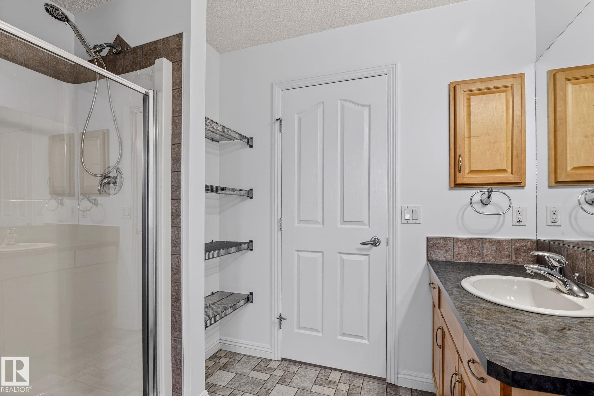 Glass-enclosed shower with tiled surround, wood-finish vanity with integrated sink, full-width mirror, built-in shelving, and paneled door - 978 Rutherford Road Sw, Edmonton, AB - Indoor Photo Showing Bathroom