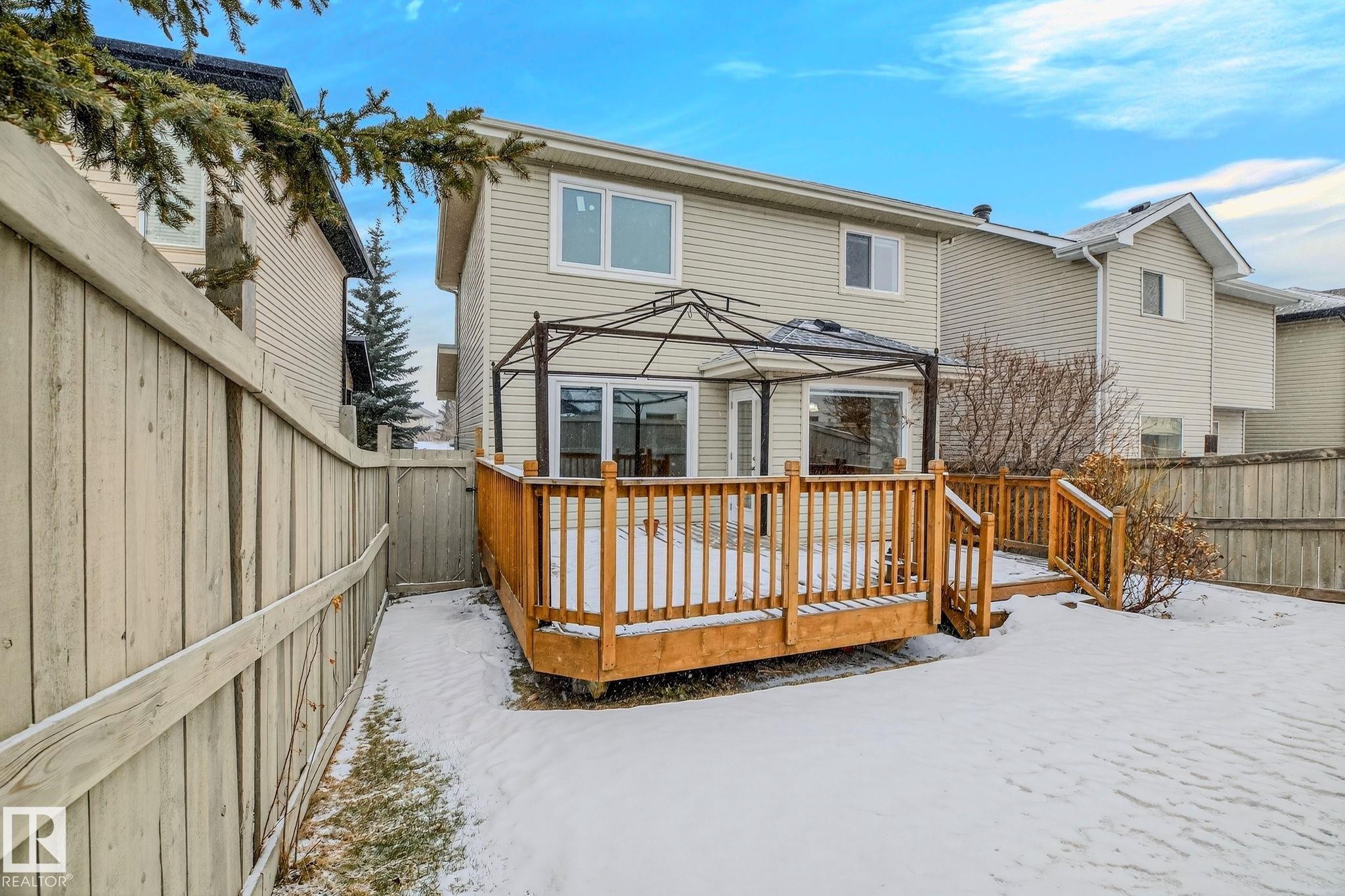 Rear exterior featuring a light-colored siding facade, a wooden deck with railing, and a framed gazebo structure - 978 Rutherford Road Sw, Edmonton, AB - Outdoor