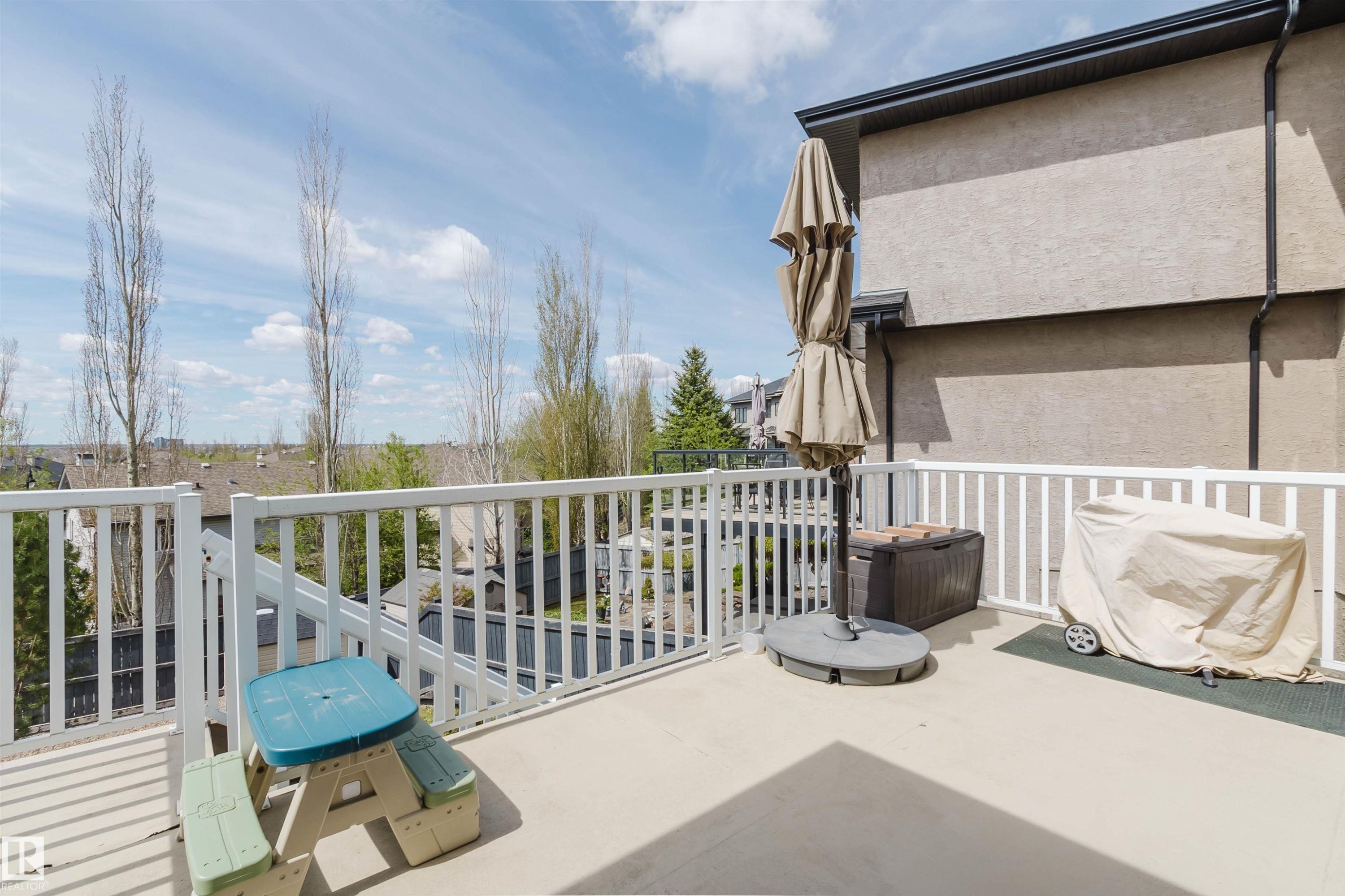 Spacious outdoor deck featuring a white railing system, stucco exterior, and a glimpse of the surrounding neighborhood rooftops and distant trees - 3412 Macneil Link, Edmonton, AB - Outdoor With Exterior
