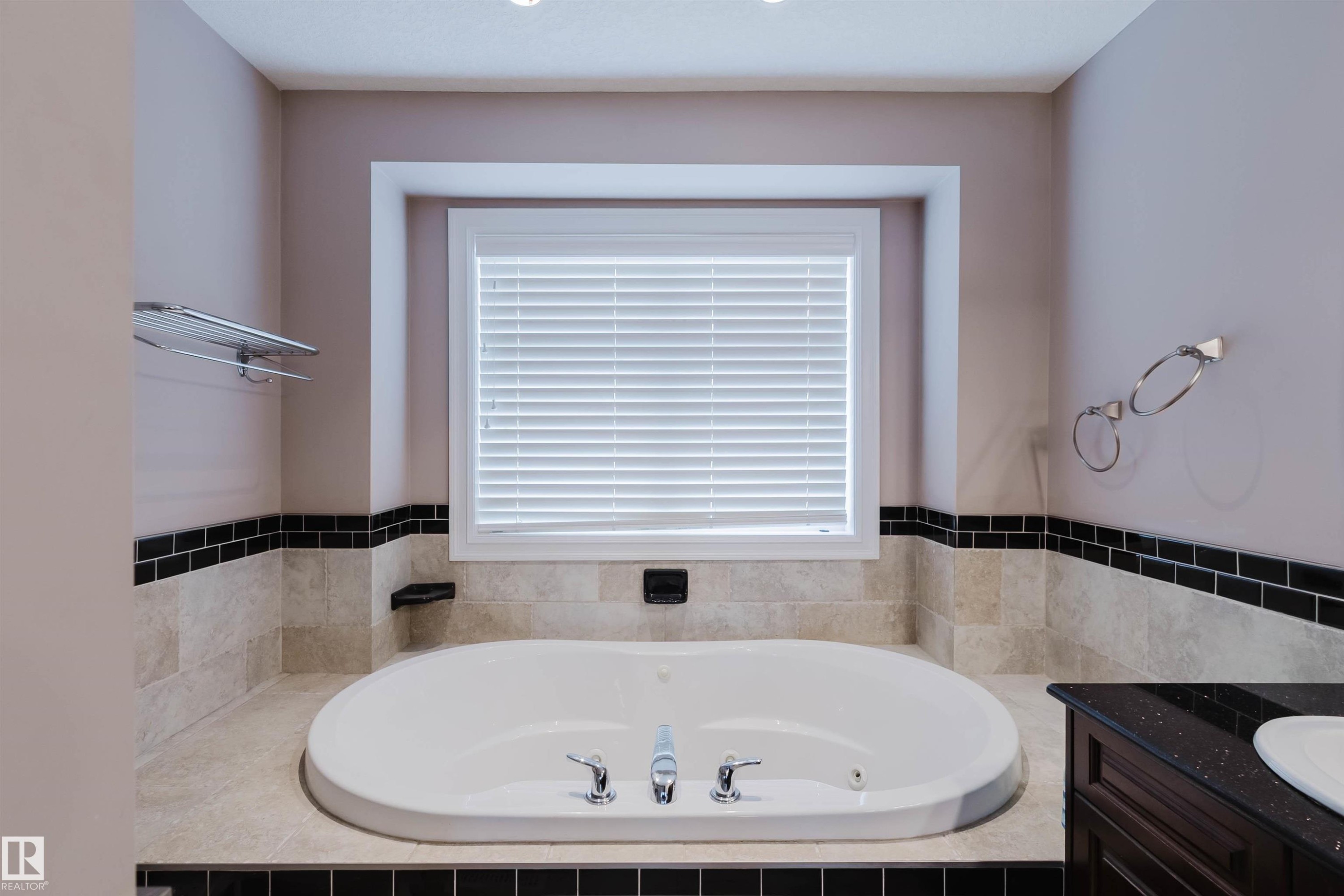 Built-in soaking tub with chrome fixtures, set within a tiled surround featuring light-toned stone and a black subway tile accent - 3412 Macneil Link, Edmonton, AB - Indoor Photo Showing Bathroom