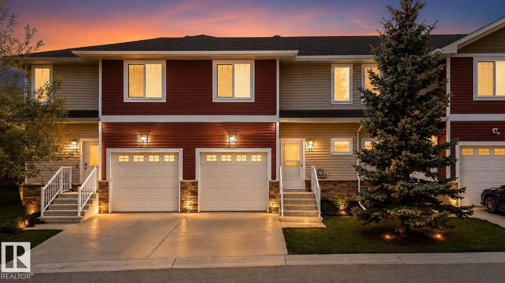 Multi-level townhome exterior featuring red and beige siding, a two-car garage with panel doors, and integrated outdoor lighting - 11 450 Mcconachie Way, Edmonton, AB - Outdoor With Facade