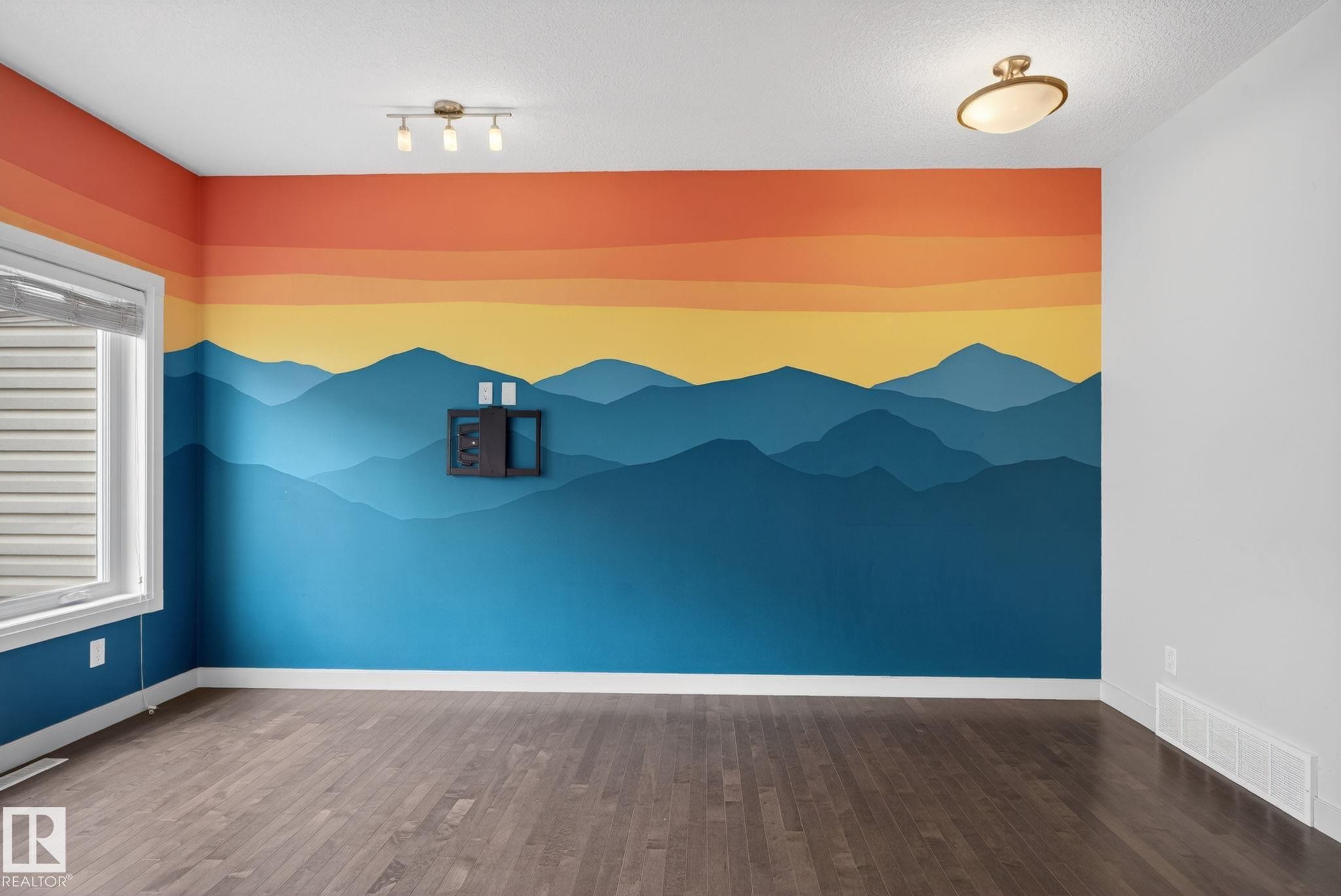 Room featuring wood-finish flooring, a vibrant mural wall, a double-pane window, and two ceiling-mounted light fixtures - 11 450 Mcconachie Way, Edmonton, AB - Indoor Photo Showing Other Room