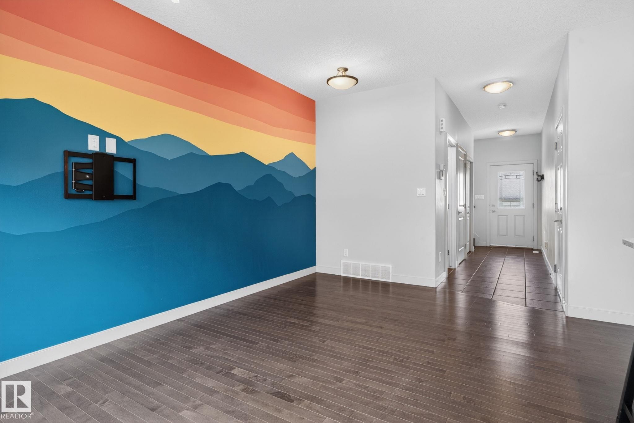Open living area featuring a decorative accent wall with a mountain mural and dark wood-finish flooring - 11 450 Mcconachie Way, Edmonton, AB - Indoor Photo Showing Other Room