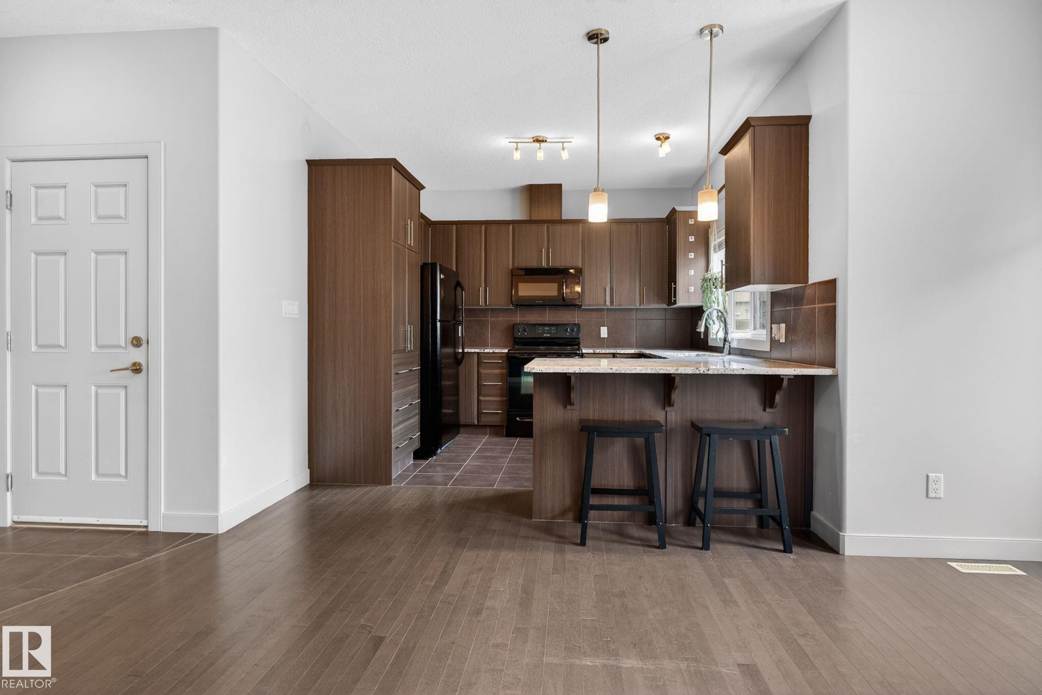 Open-concept kitchen featuring wood-finish cabinetry, black appliances, and a breakfast bar with pendant lighting - 11 450 Mcconachie Way, Edmonton, AB - Indoor Photo Showing Kitchen