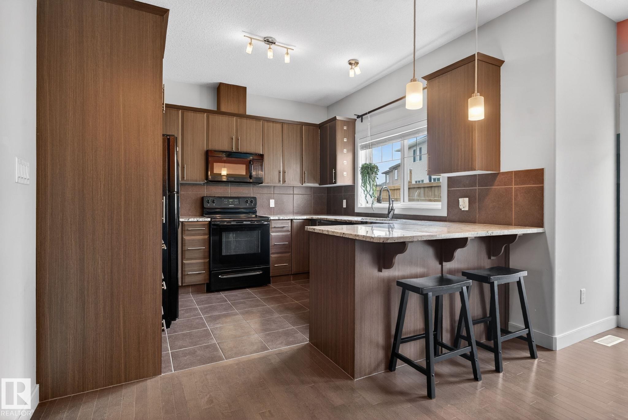 Kitchen featuring wood-finish cabinetry, black appliances, a tile backsplash, and a breakfast bar with stone-finish countertops - 11 450 Mcconachie Way, Edmonton, AB - Indoor Photo Showing Kitchen