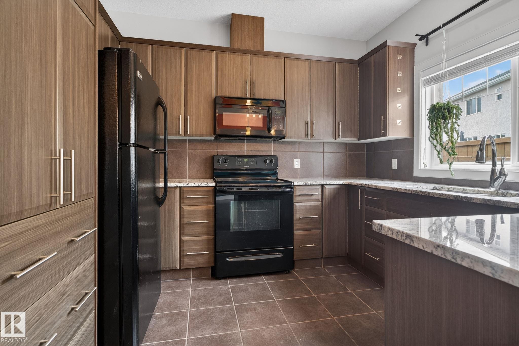 Kitchen featuring wood-finish cabinetry, black appliances, stone-look countertops, a tile backsplash, and tile flooring - 11 450 Mcconachie Way, Edmonton, AB - Indoor Photo Showing Kitchen