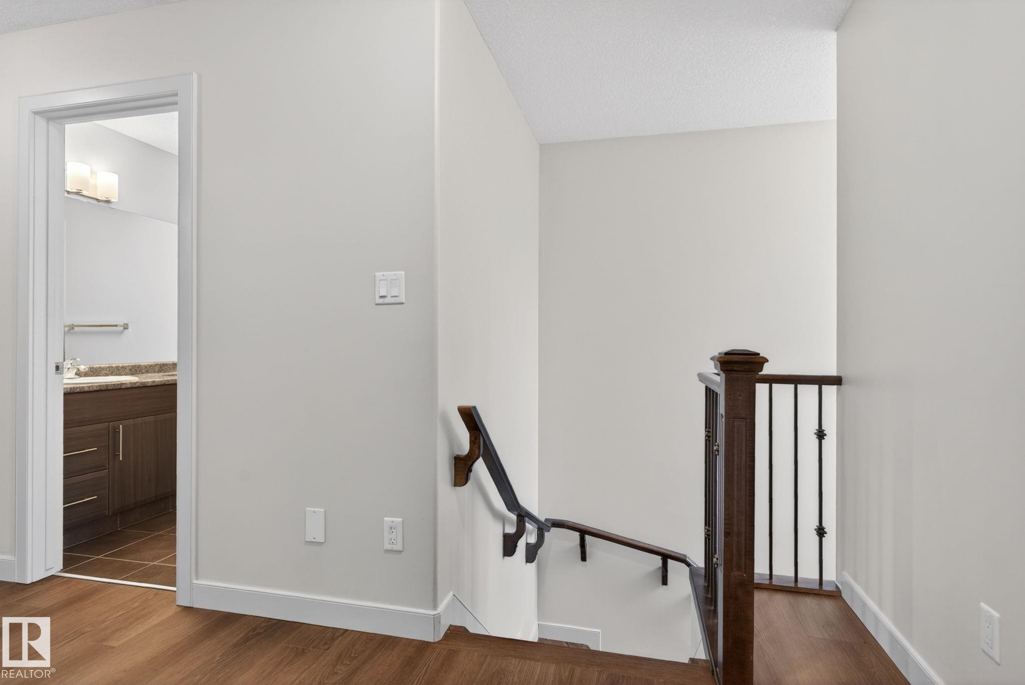 Hardwood-finish flooring extending from the hallway into the bathroom, featuring a dark wood newel post and black metal balusters - 11 450 Mcconachie Way, Edmonton, AB - Indoor Photo Showing Other Room