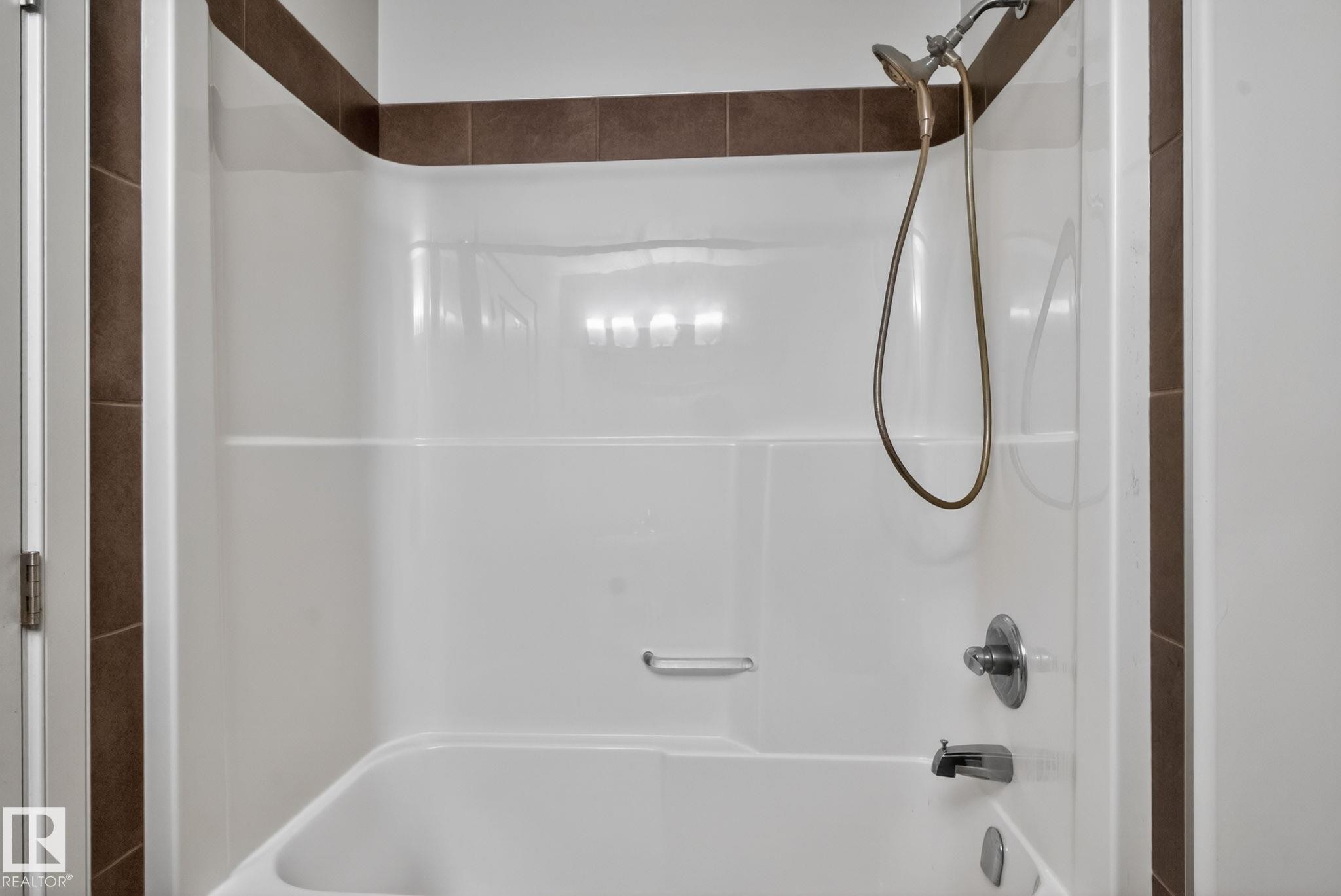 White bathtub and shower combination unit featuring a handheld showerhead with a bronze-finish hose and chrome fixtures - 11 450 Mcconachie Way, Edmonton, AB - Indoor Photo Showing Bathroom