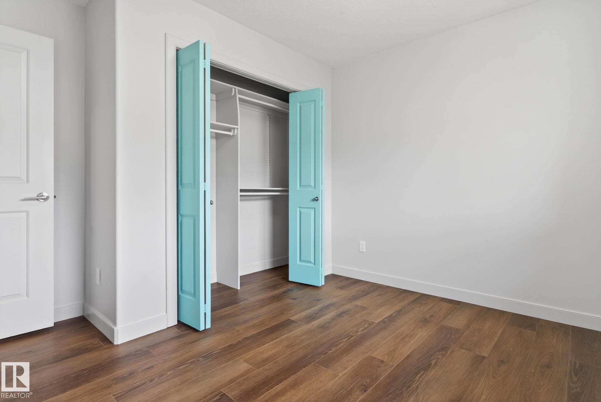 Bedroom featuring warm wood-finish flooring, white baseboards, a white interior door with a chrome handle, white walls, and a closet with bifold turquoise doors - 11 450 Mcconachie Way, Edmonton, AB - Indoor Photo Showing Other Room
