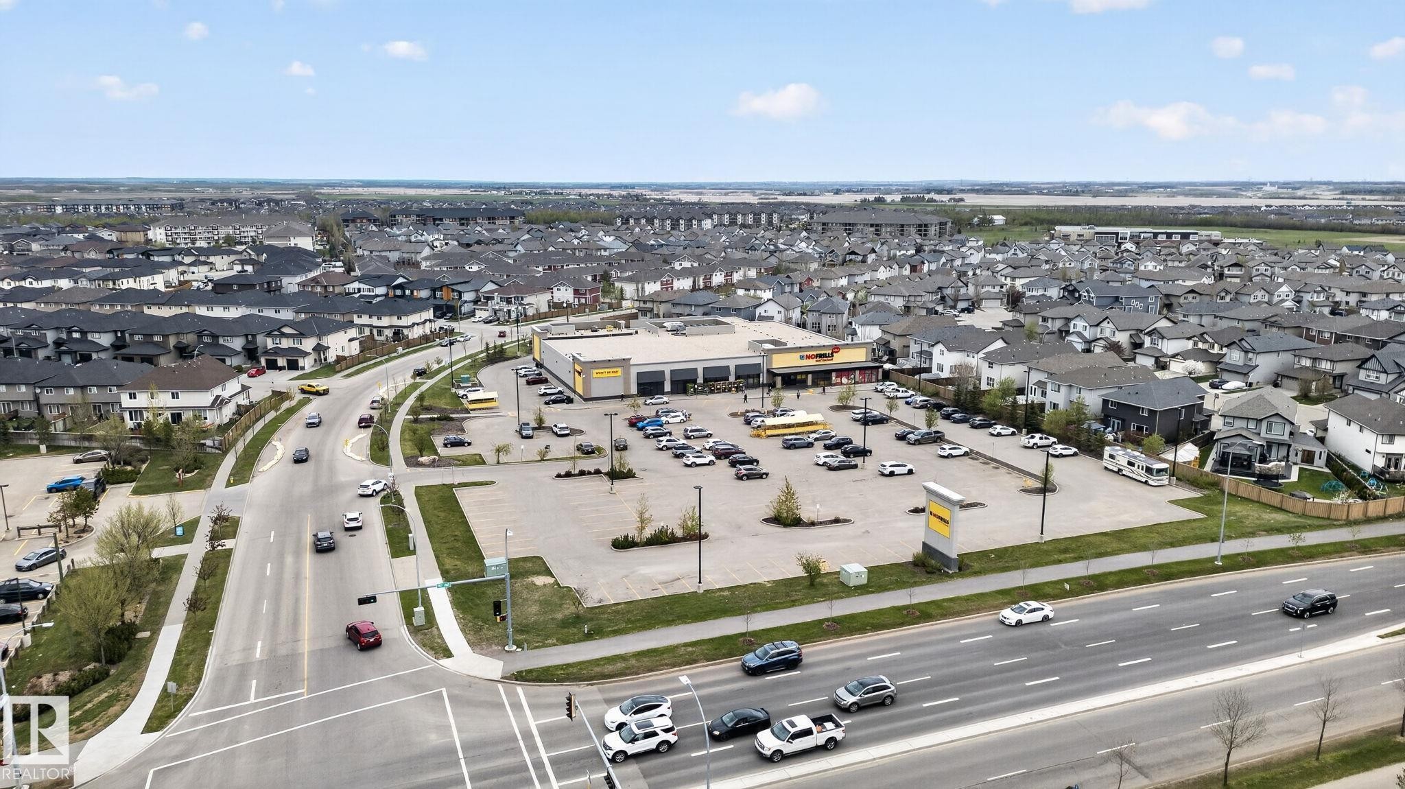 Aerial overview of a suburban community featuring a commercial center with ample parking, surrounded by residential properties with dark-pitched roofs - 11 450 Mcconachie Way, Edmonton, AB - Outdoor With View