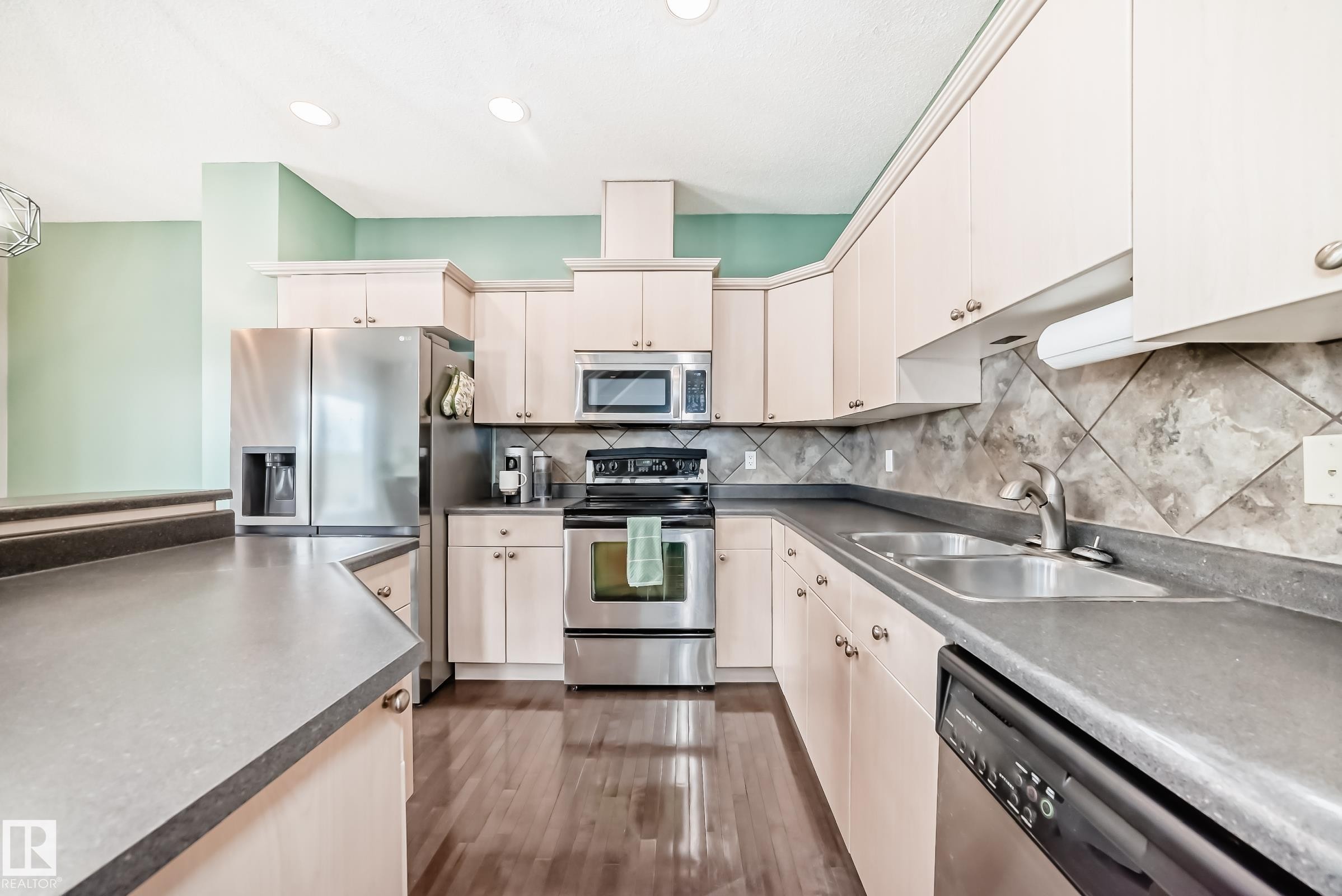 Spacious kitchen featuring light-toned cabinetry, stainless steel appliances, dark countertops, and wood-finish flooring - 105 13825 155 Avenue, Edmonton, AB - Indoor Photo Showing Kitchen With Stainless Steel Kitchen With Double Sink
