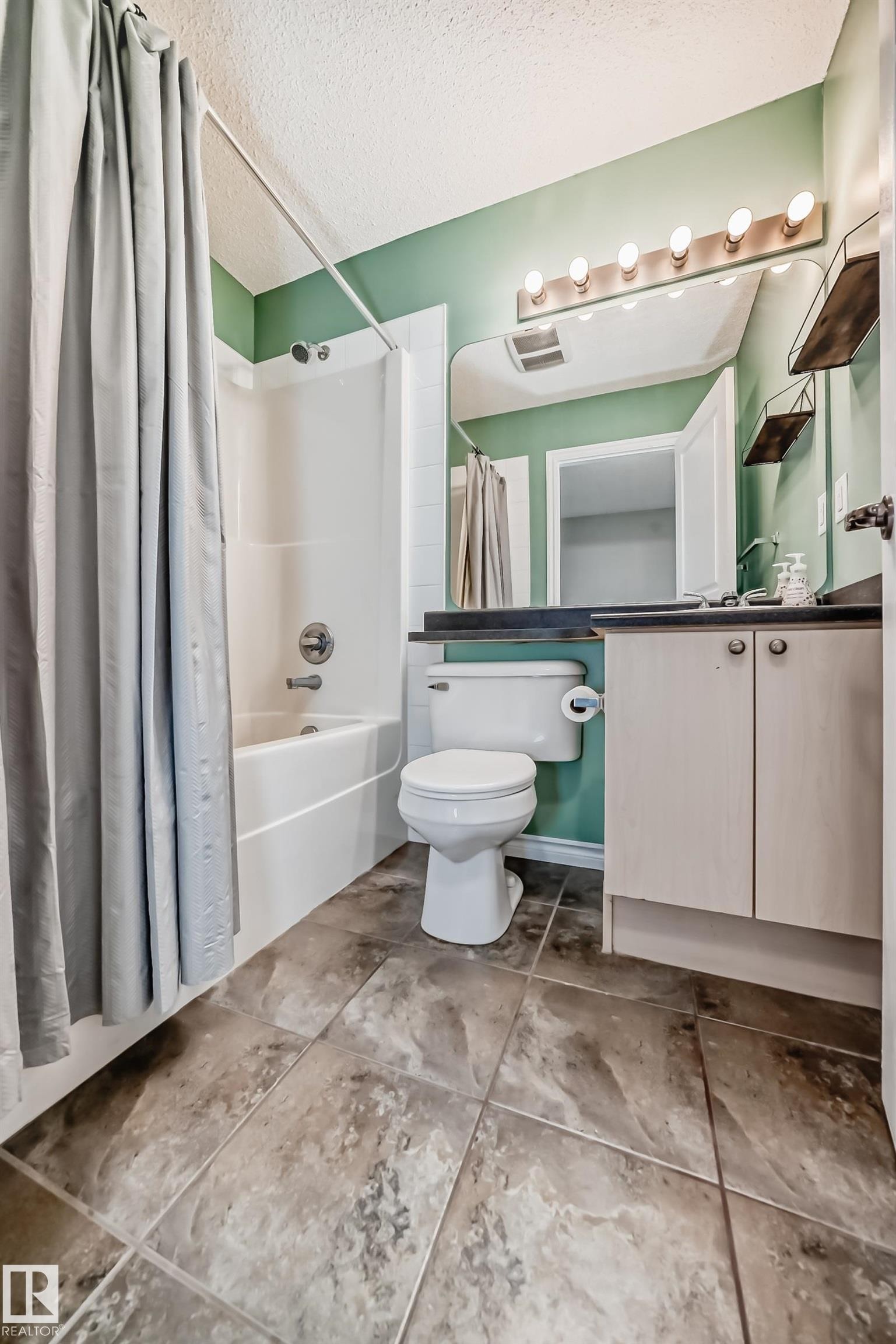 Bathroom featuring a white bathtub/shower combination, a wall-mounted vanity light fixture, a rectangular mirror, a white toilet, and a light-colored vanity with a dark countertop - 105 13825 155 Avenue, Edmonton, AB - Indoor Photo Showing Bathroom