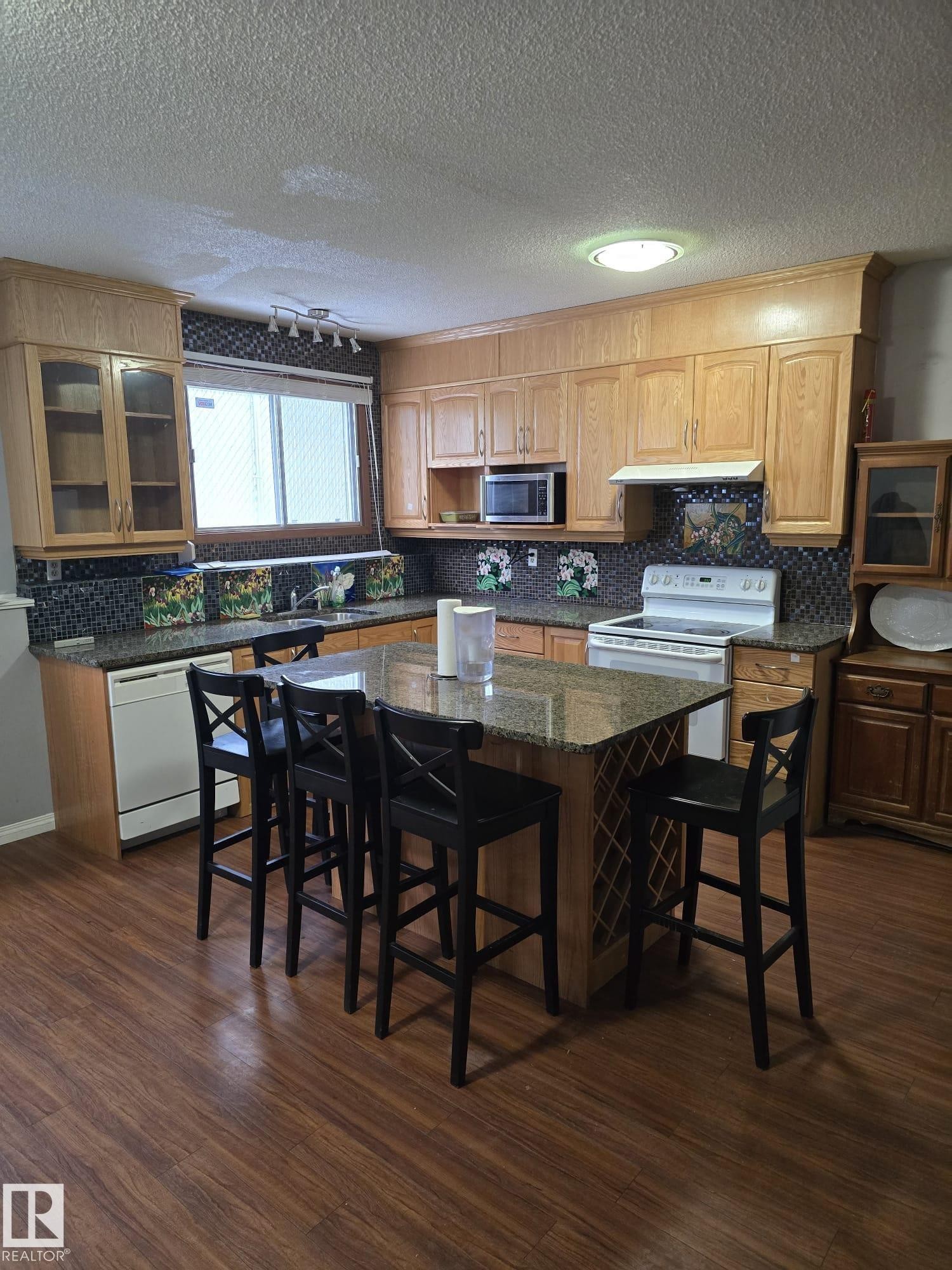 Kitchen featuring extensive light wood cabinetry, granite-finish countertops, tile backsplash, wood-finish flooring, and a central island with integrated wine storage - 11927 85 Street Nw, Edmonton, AB - Indoor
