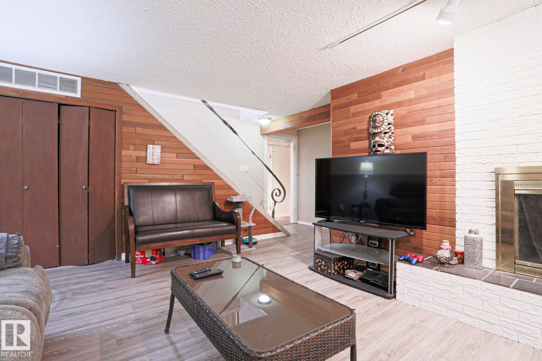 Living area featuring wood-finish flooring, a white brick fireplace with a brass-finish insert, wood panel accent walls, and bi-fold closet doors - 1214 85 Street Nw, Edmonton, AB - Indoor Photo Showing Living Room
