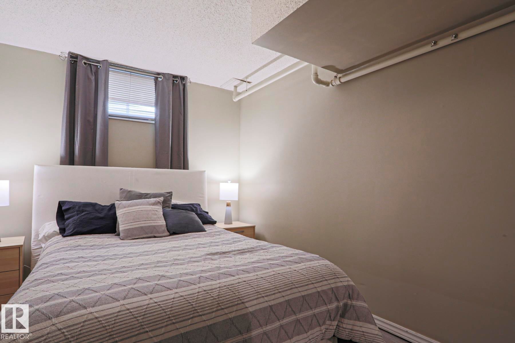 Neutral-toned room featuring a window with blinds and dark gray curtains, a white upholstered headboard, and light wood-finish nightstands with lamps - 1214 85 Street Nw, Edmonton, AB - Indoor Photo Showing Bedroom