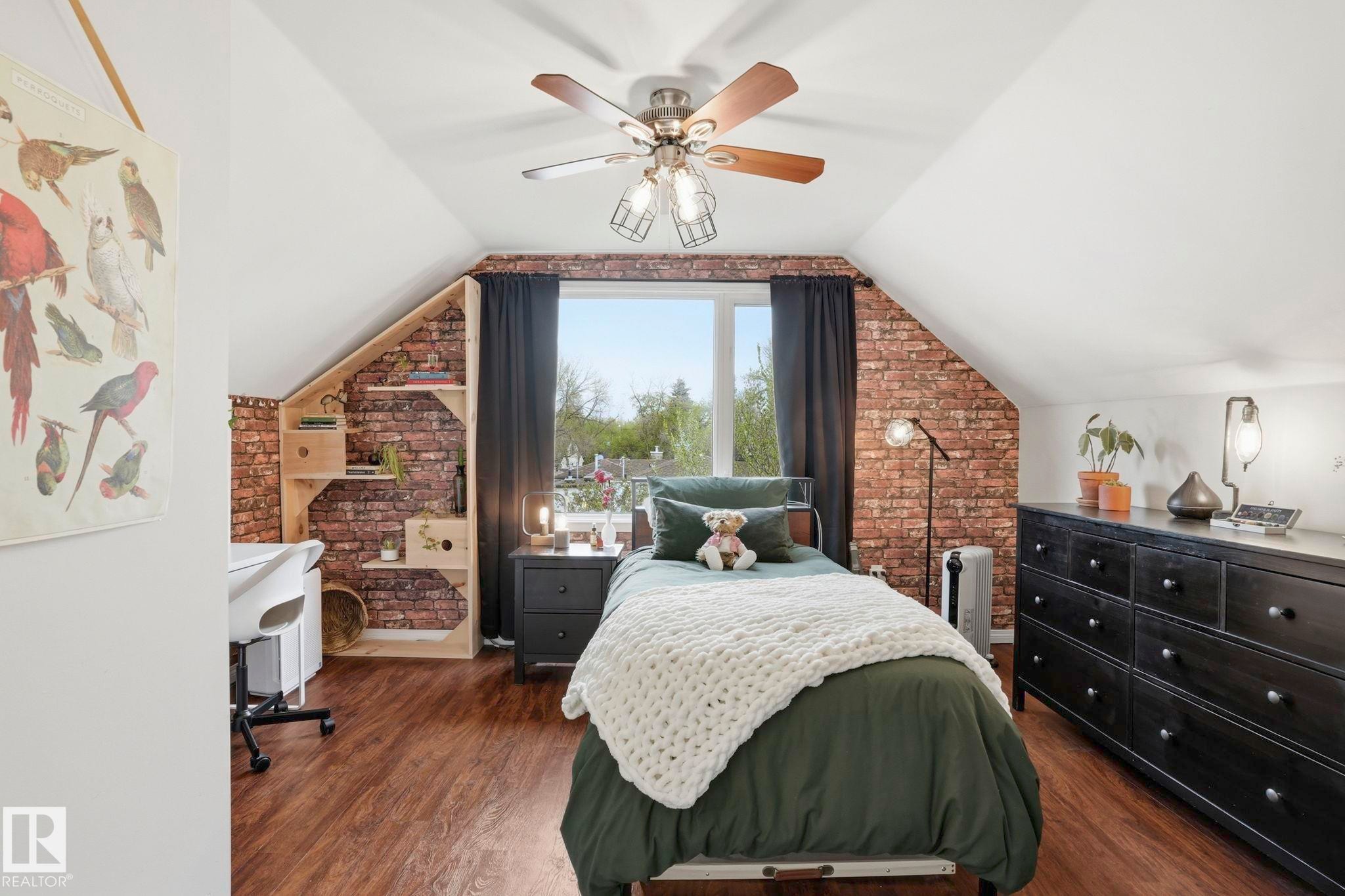 Attic room featuring wood-finish flooring, an exposed brick accent wall, built-in shelving, a ceiling fan with light fixture, and a large window - 4 Langley Dr, Fort Saskatchewan, AB - Indoor Photo Showing Bedroom
