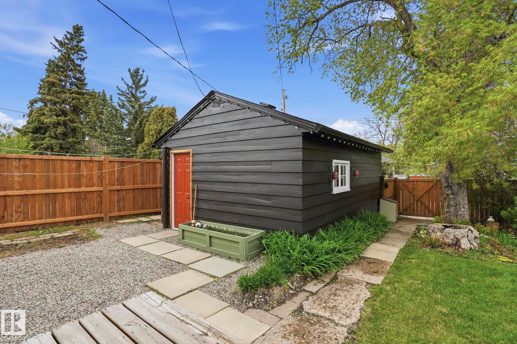 Detached outbuilding with dark siding and a red door, featuring a white-trimmed window, concrete paver pathway, and a wooden fence - 4 Langley Dr, Fort Saskatchewan, AB - Outdoor