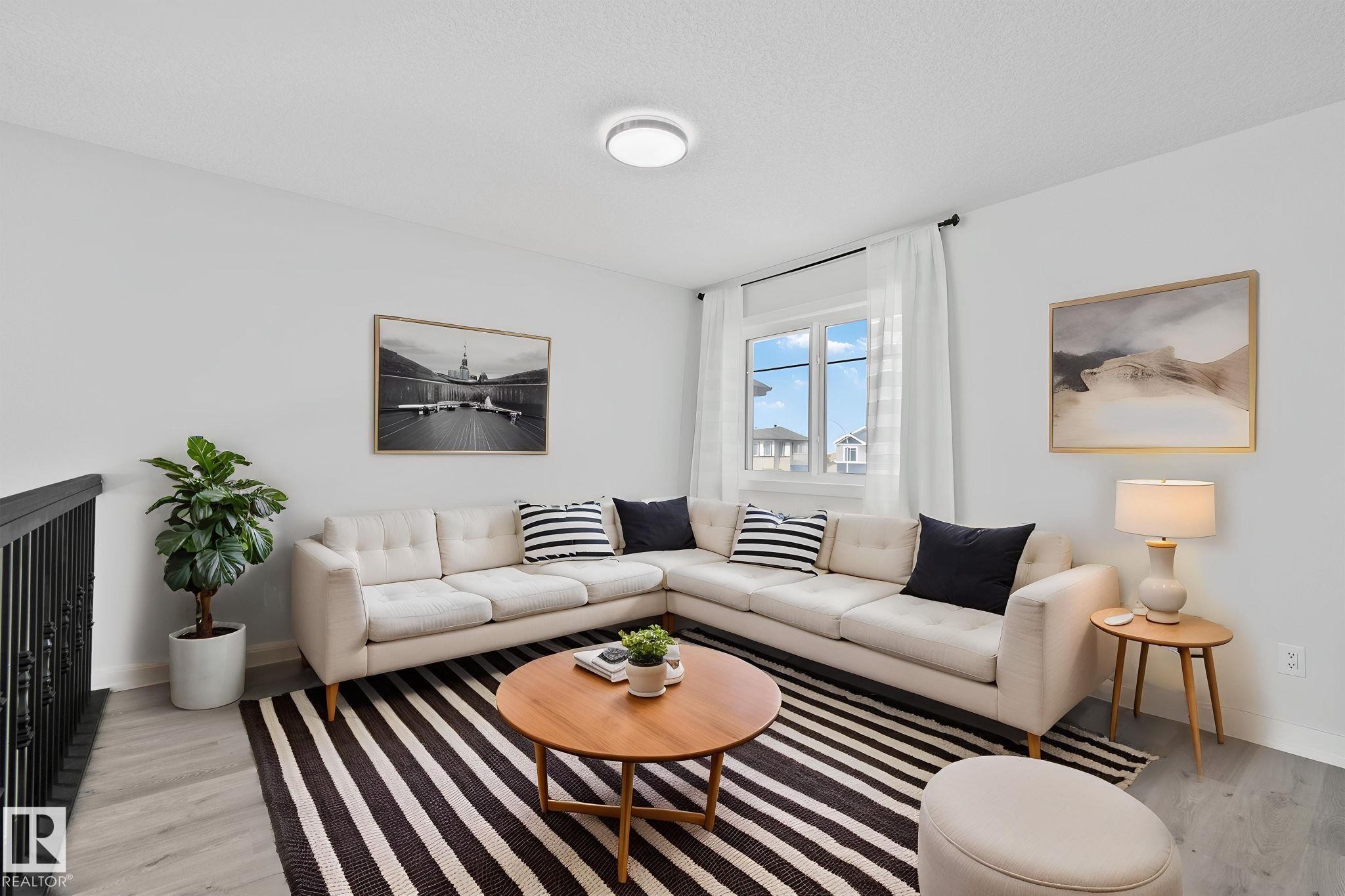 Living area featuring wood-finish flooring, a contemporary ceiling light fixture, a window with white curtains, and a black wrought iron railing - 334 Aston Close, Leduc, AB - Indoor Photo Showing Living Room